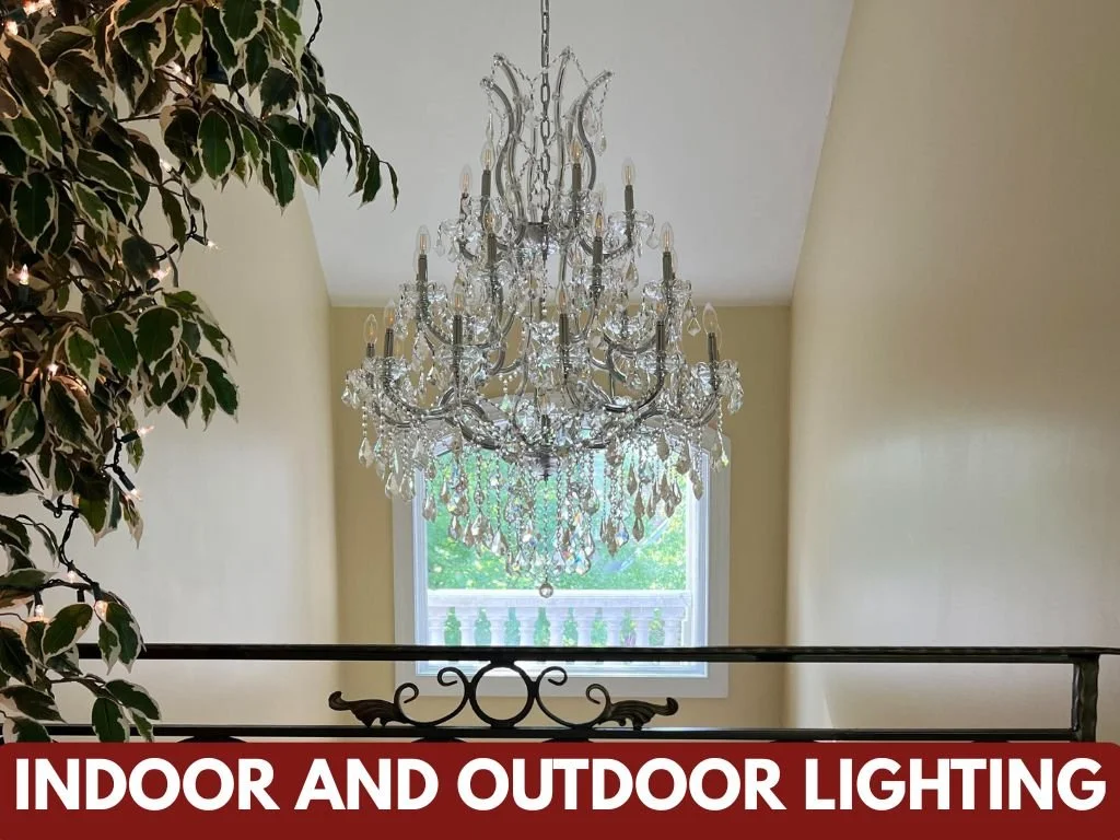 A large, ornate crystal chandelier hanging from the ceiling in front of a window with a view of greenery outside, and a potted plant partially visible on the left side.