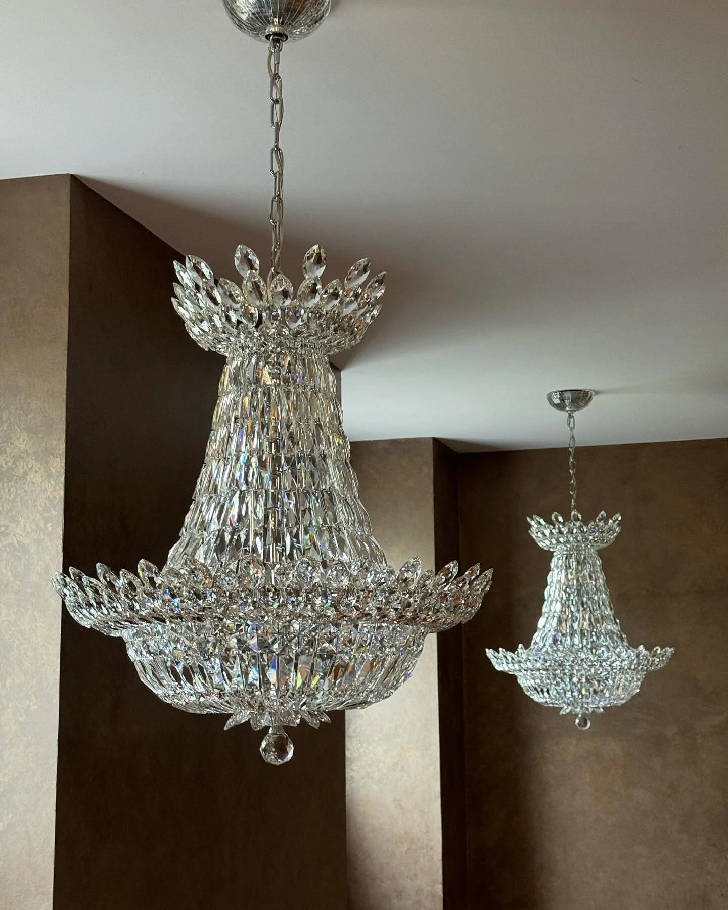 Two ornate crystal chandeliers hanging from a ceiling in a room with dark brown walls.