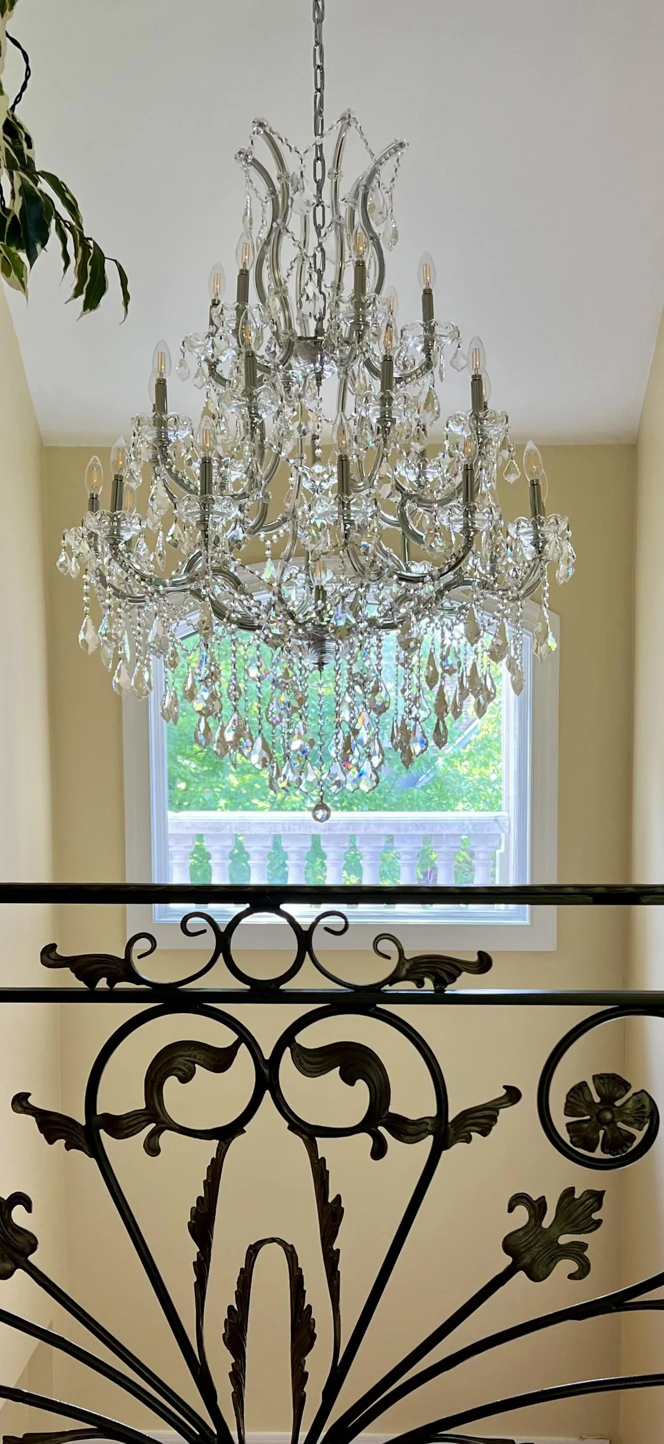 A large, ornate crystal chandelier hanging from the ceiling in a room with a window and yellow walls. The chandelier has multiple candle-style lights and hanging crystal prisms.