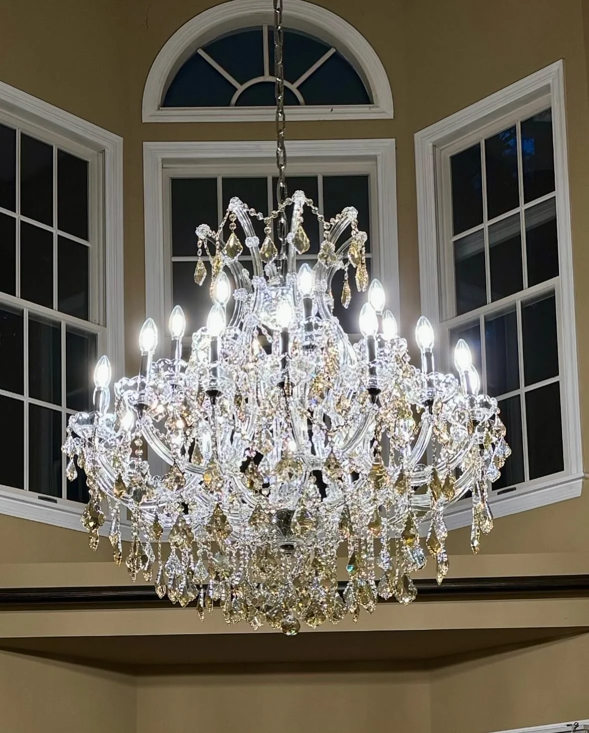 A large, ornate crystal chandelier hanging from the ceiling with multiple lit bulbs, surrounded by tall arched windows with double-pane glass.