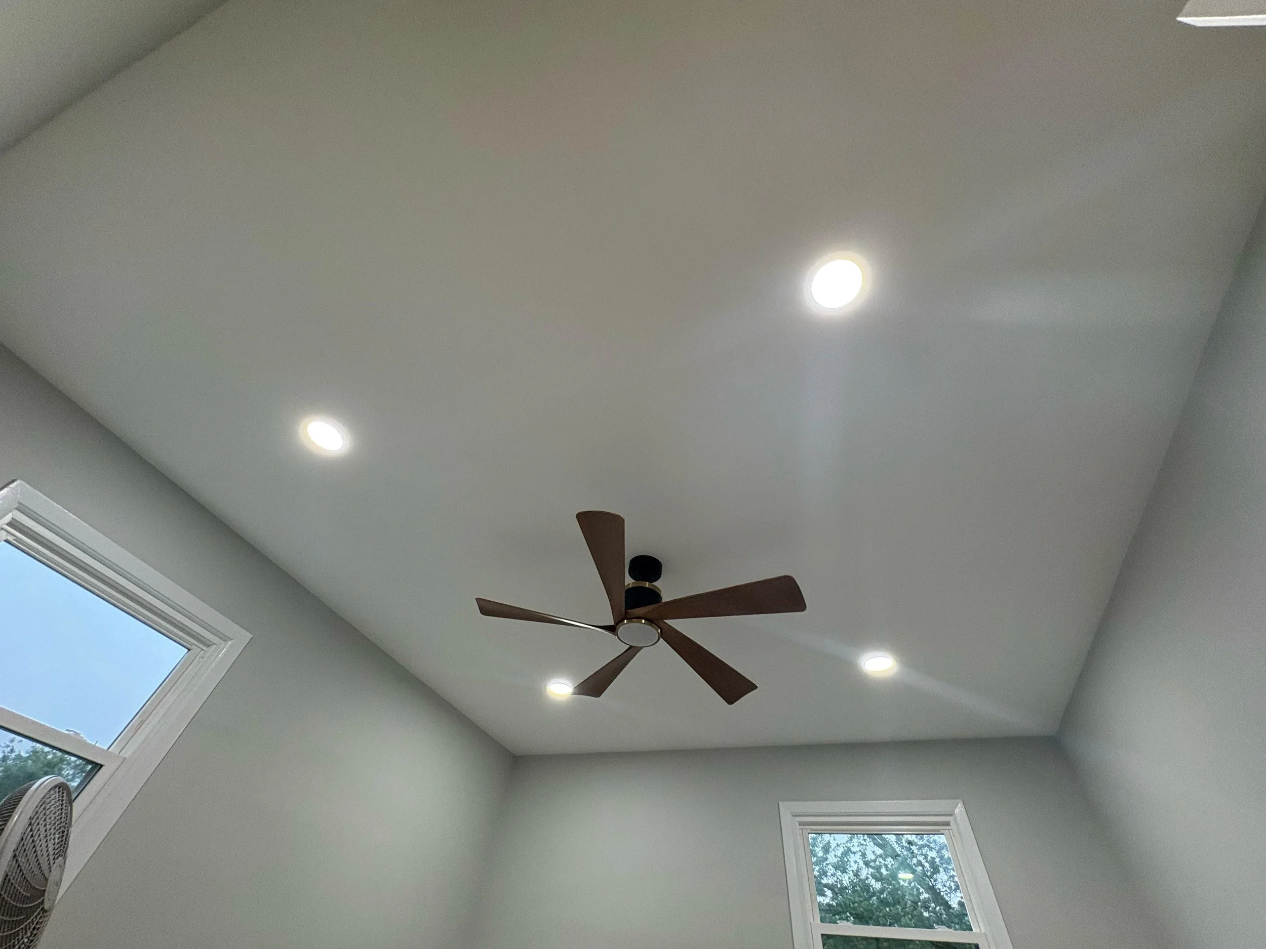 Ceiling with recessed lighting, a ceiling fan with brown blades, two windows with views of trees, and light gray walls.