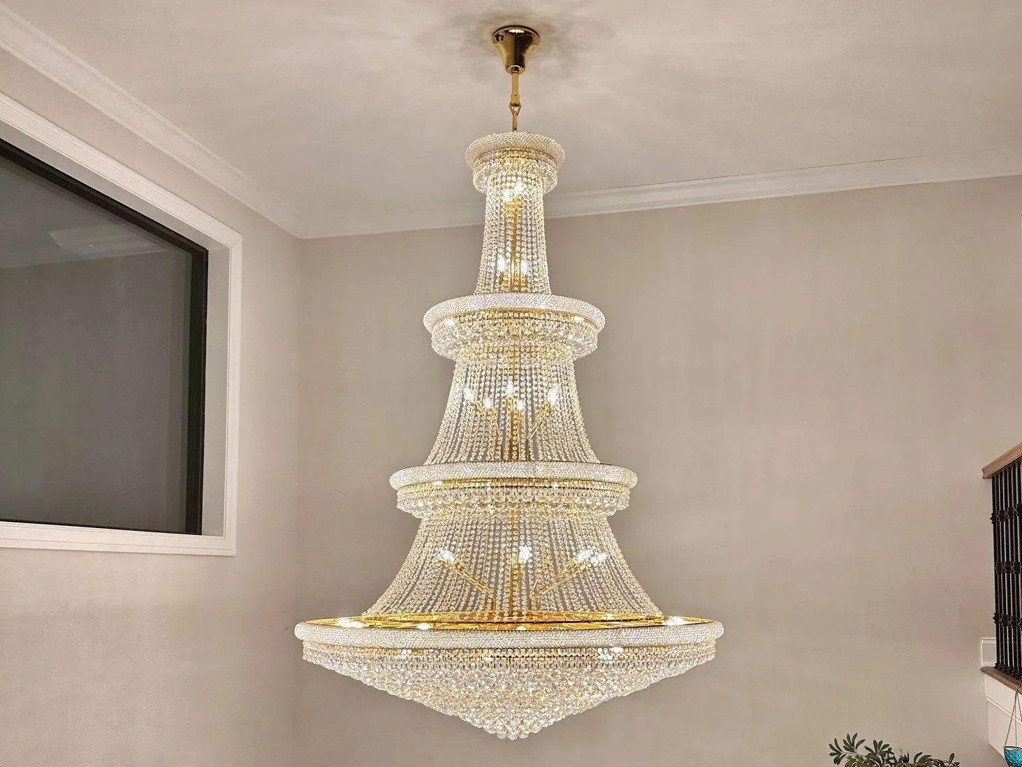 A large, ornate chandelier hanging from the ceiling in a room with beige walls and a window on the left side.
