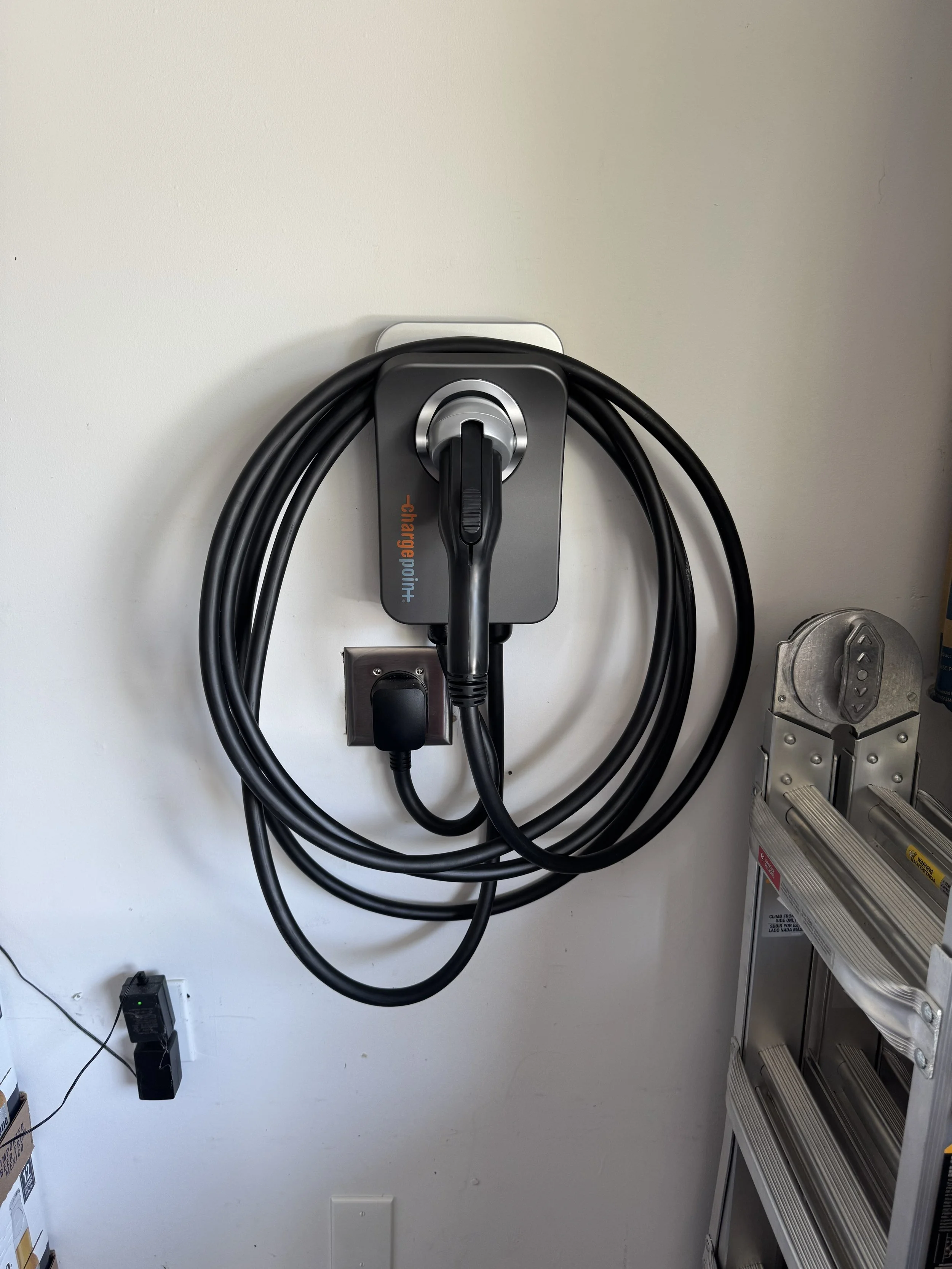 Electric vehicle charging station mounted on a wall with charging cable plugged in, next to a metal ladder and an electrical outlet with a black device plugged into it.