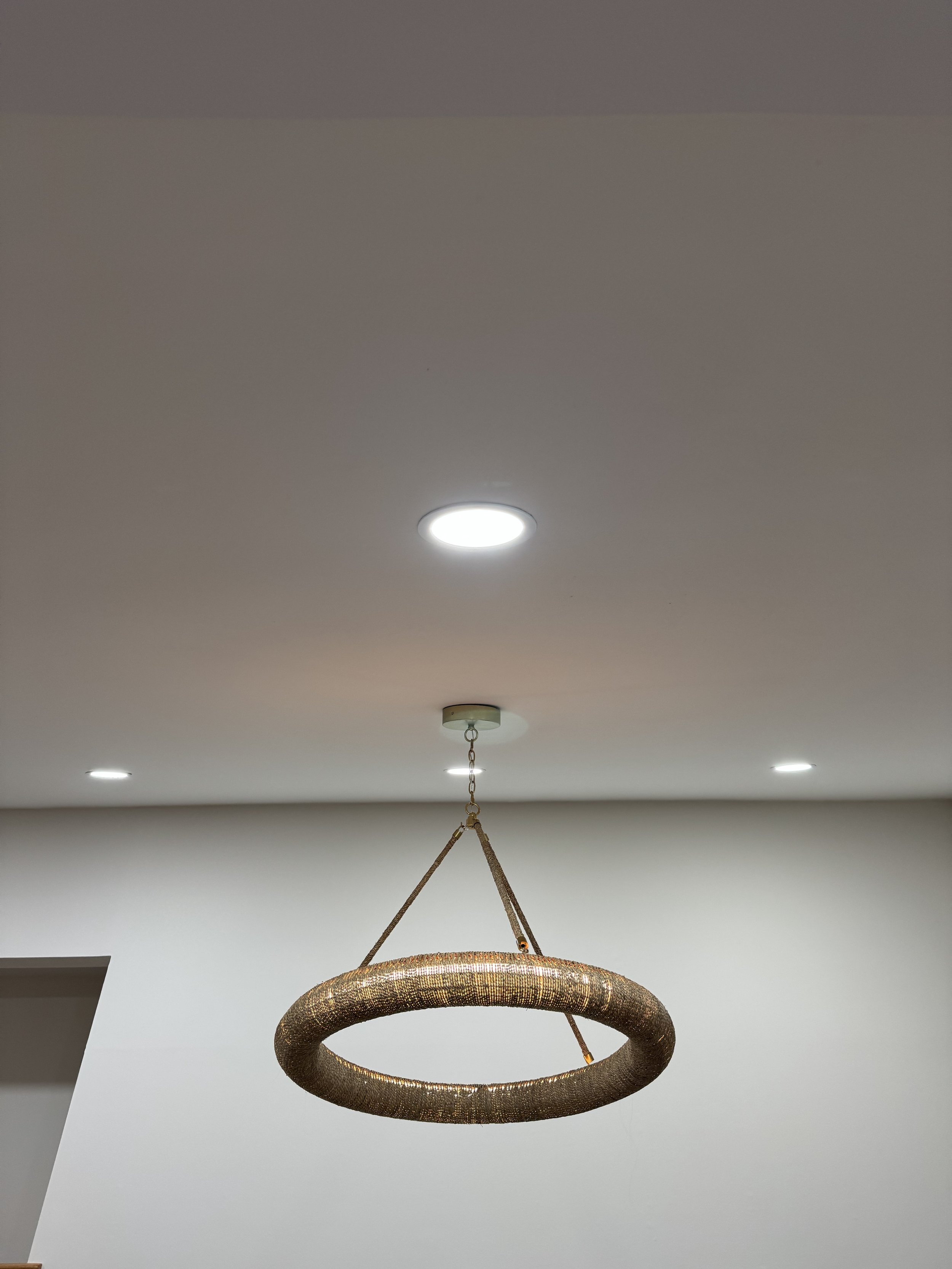 A modern gold circular chandelier hanging from a chain from a plain white ceiling with recessed lighting.