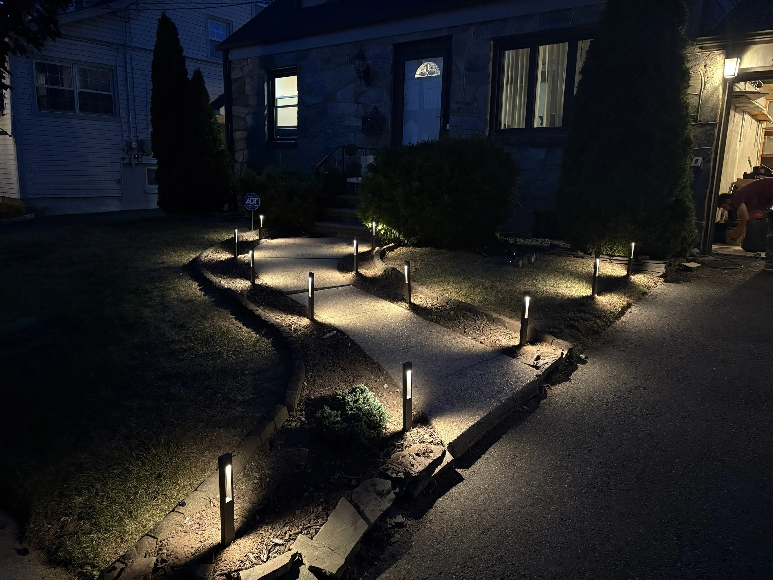 Front yard of house with a curved concrete walkway illuminated by small garden lights, flanked by bushes and a lawn, during early evening or night.
