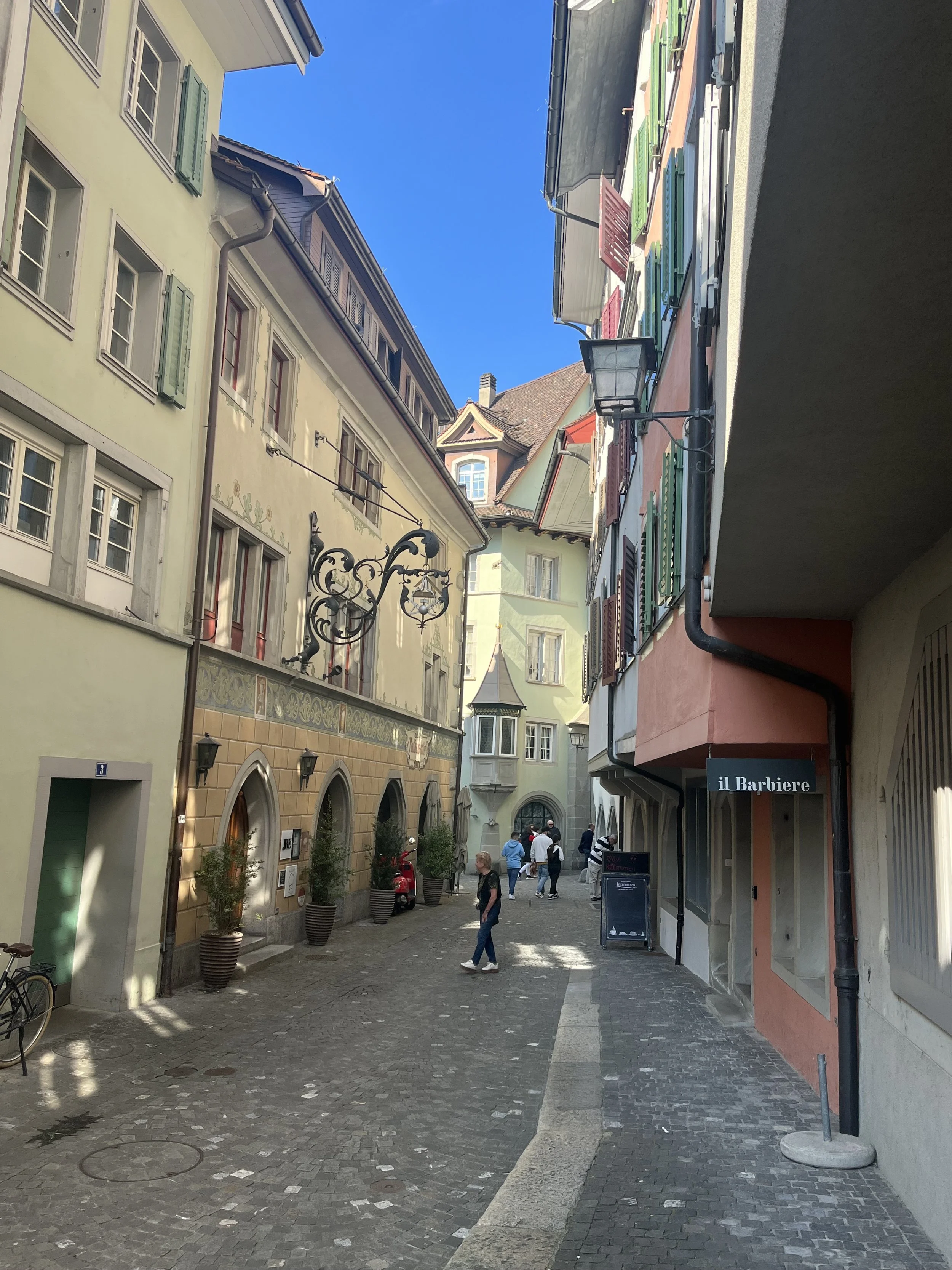 A narrow European street with colorful buildings, potted plants, and a few pedestrians walking. Clear blue sky overhead.