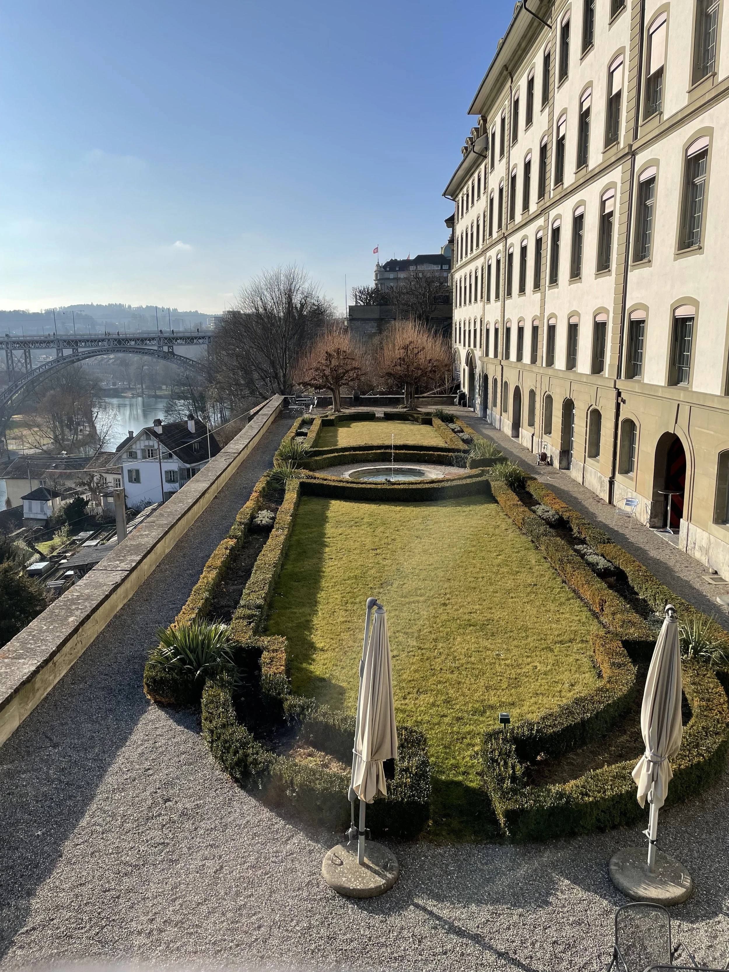 A neatly manicured garden with a fountain at the center, surrounded by a low hedge, with two closed umbrellas in the foreground and a large building with multiple windows on the right side. A bridge spans across a river in the background under a clear blue sky.