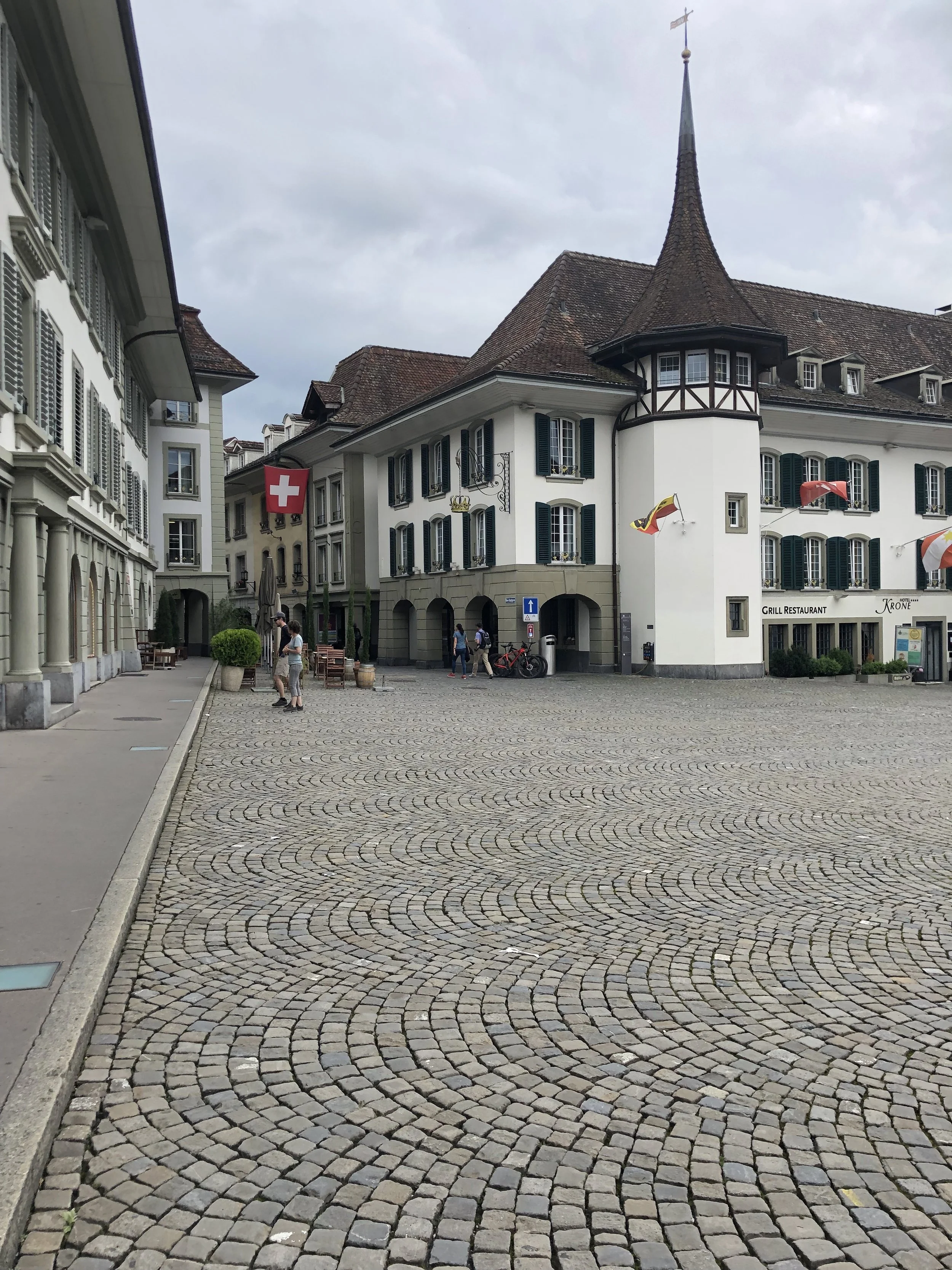 A European town square with cobblestone pavement, historic buildings with dark green shutters, and flags including a Swiss flag and a flag with a yellow and red emblem. People are walking around, and there are outdoor seating areas.