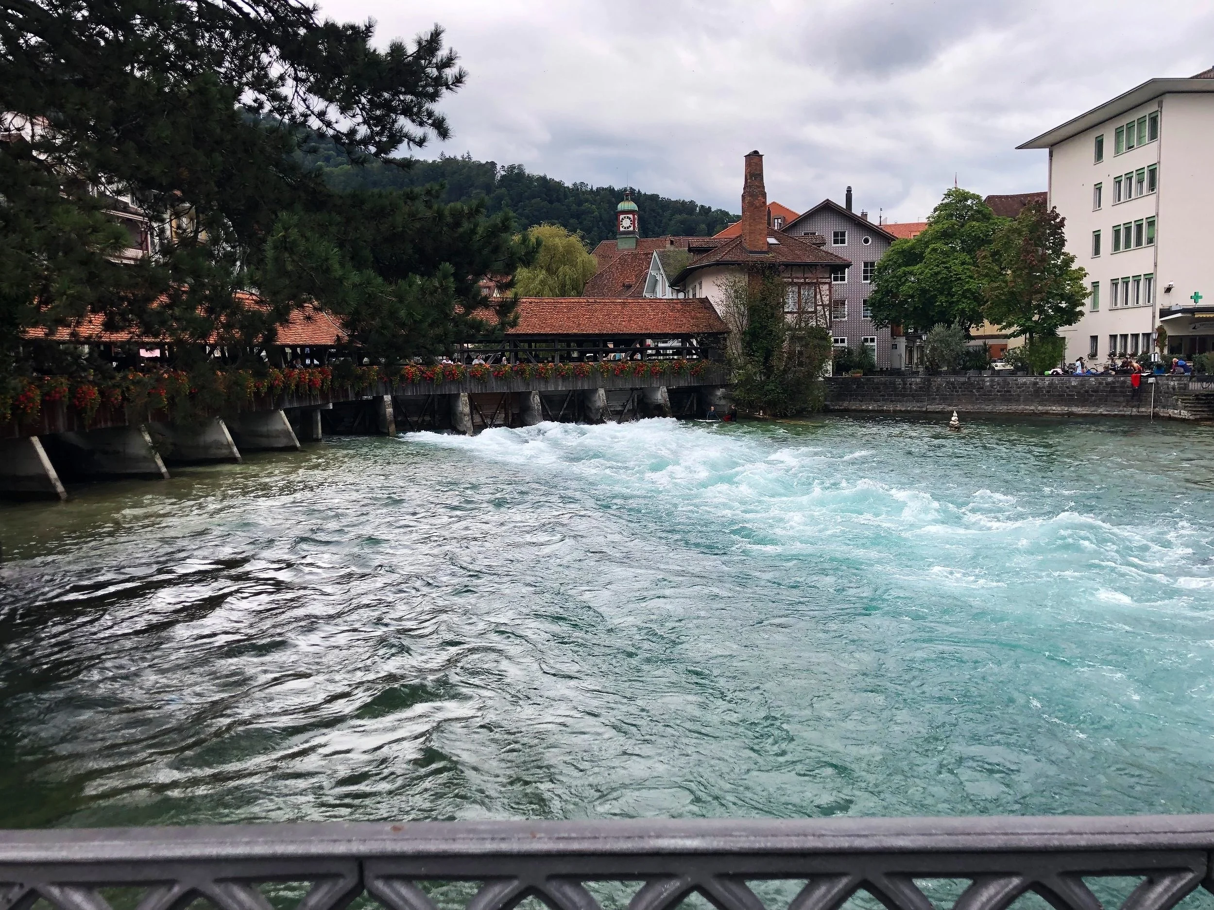 A river flowing through a town with buildings and trees along the banks, a bridge over the water, and a cloudy sky above.