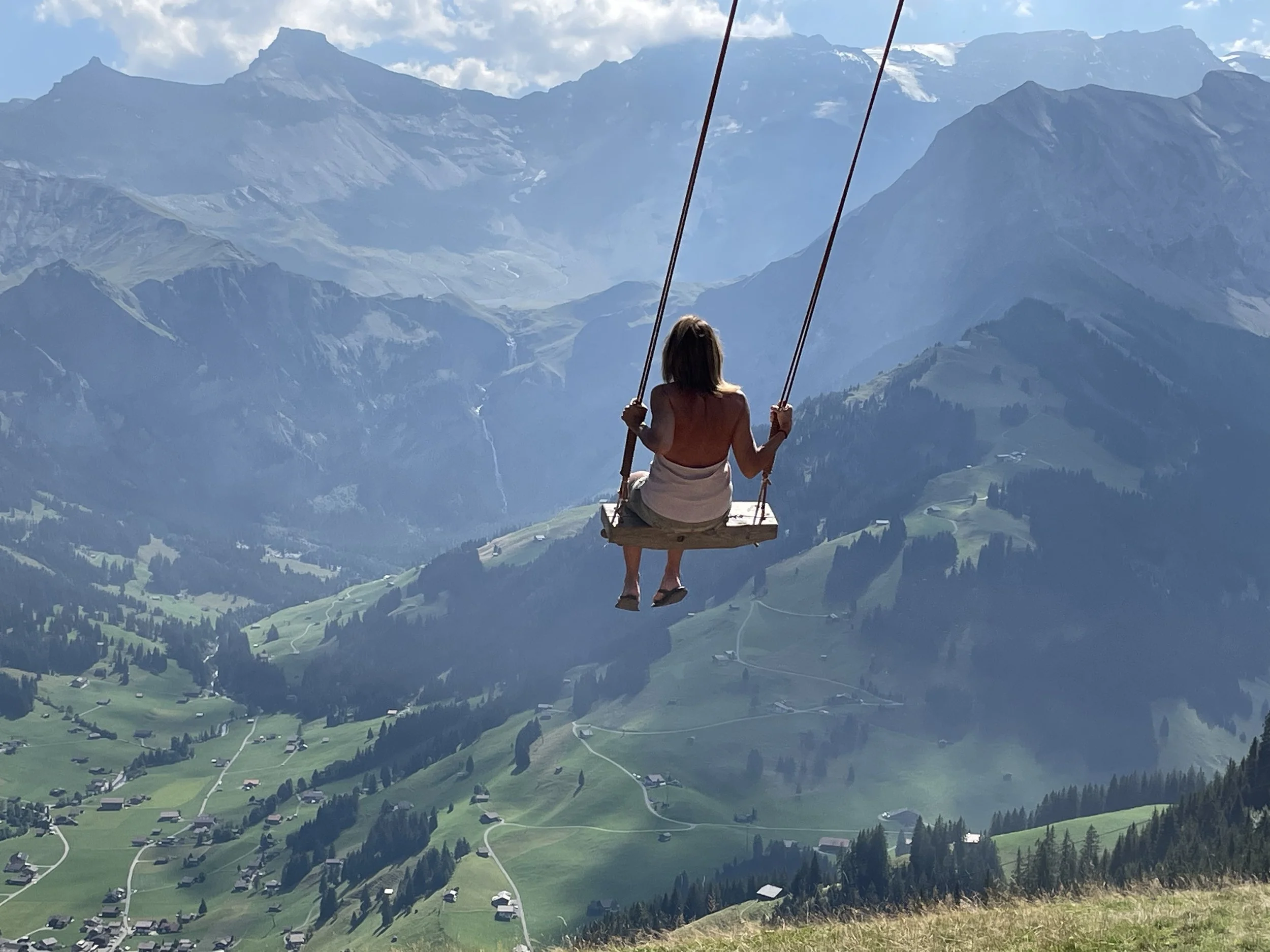 A woman sitting on a wooden swing overlooking a scenic mountain landscape with green valleys, trees, and distant peaks.