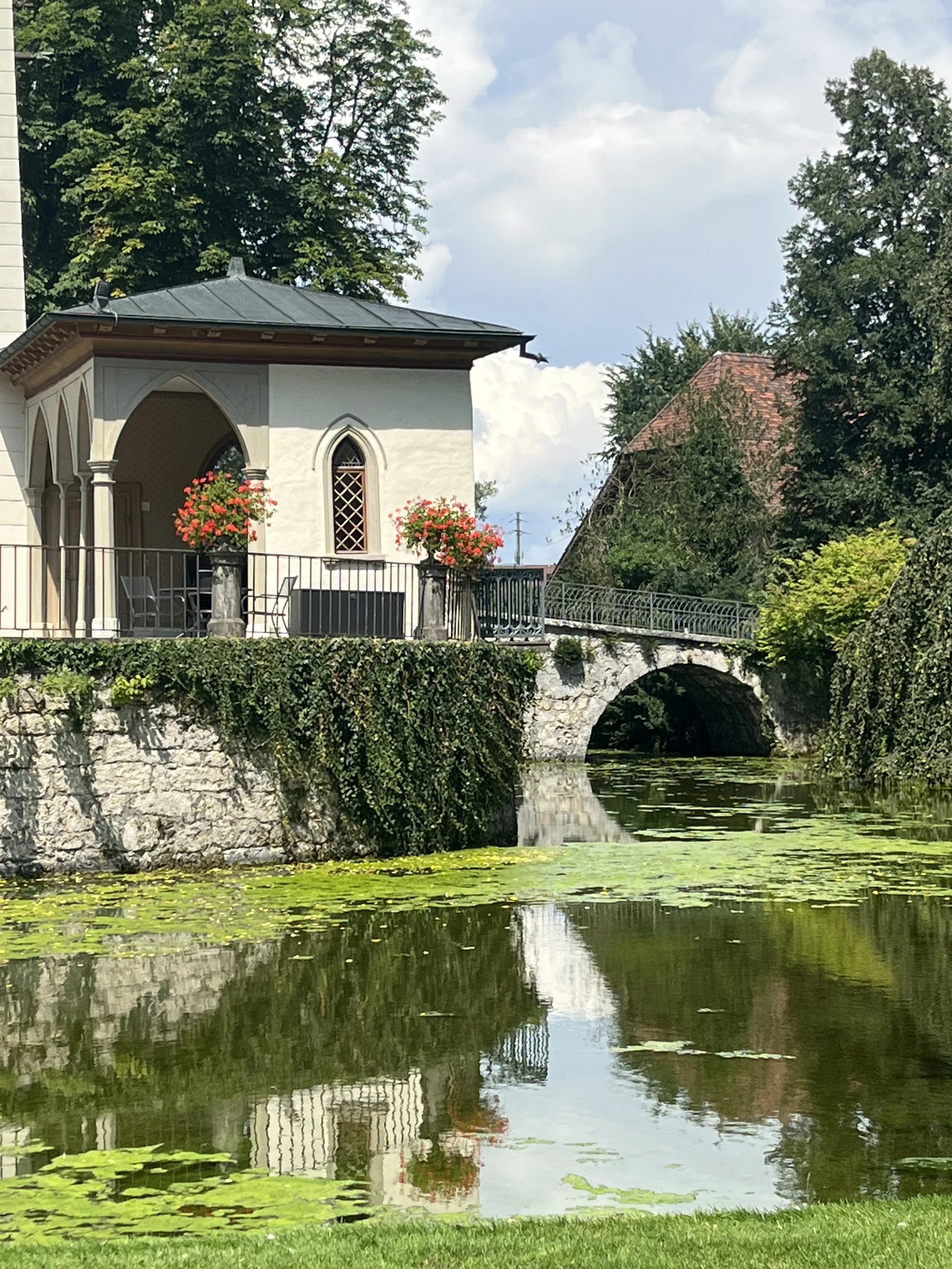 A scenic view of a small, white building with arched windows and a balcony with flower baskets, situated near a stone bridge over a pond with water lilies, surrounded by lush green trees under a partly cloudy sky.
