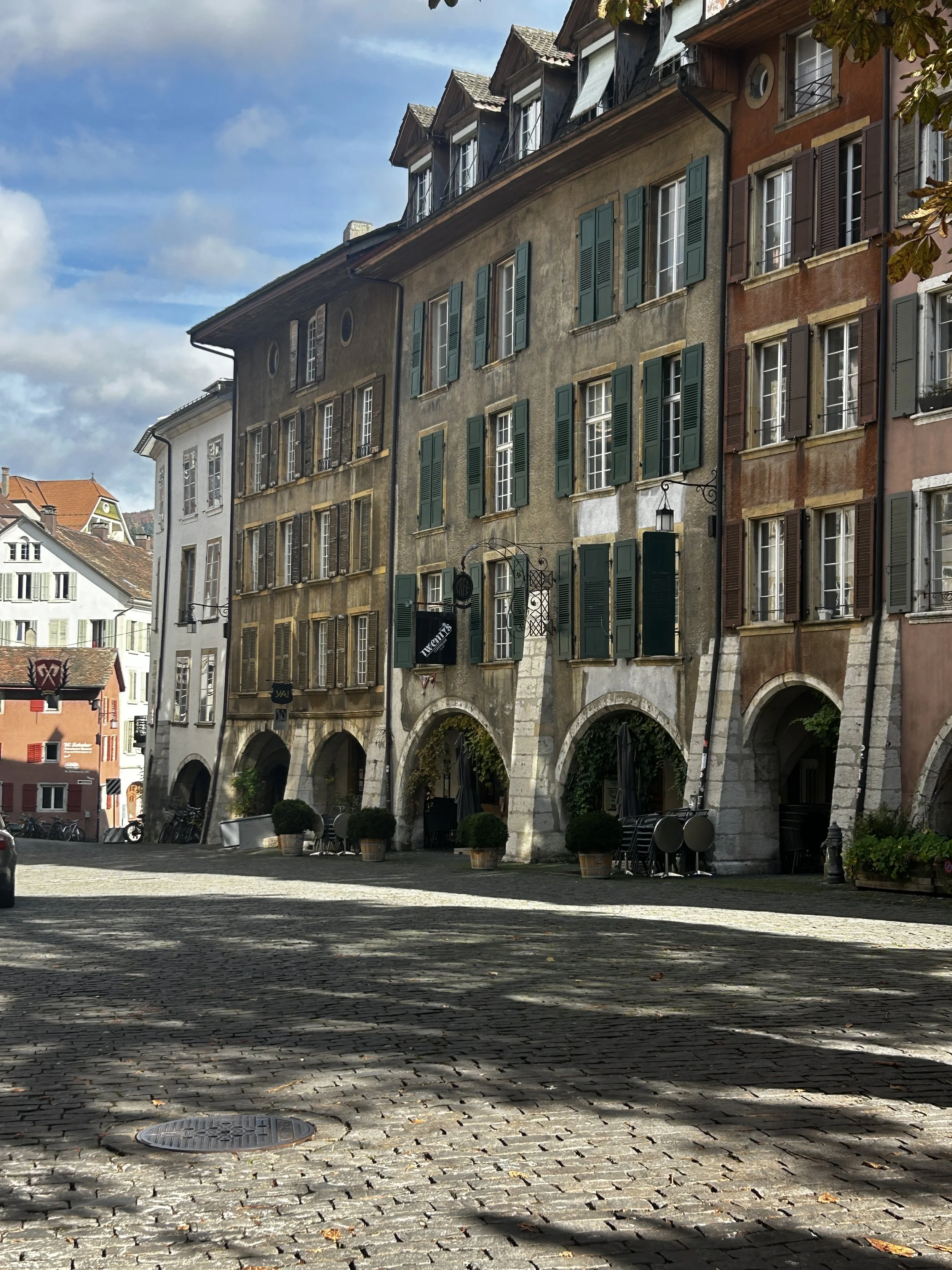 Colorful old European building with arched ground floor entrances, green and brown shutters, and outdoor tables on cobblestone street.