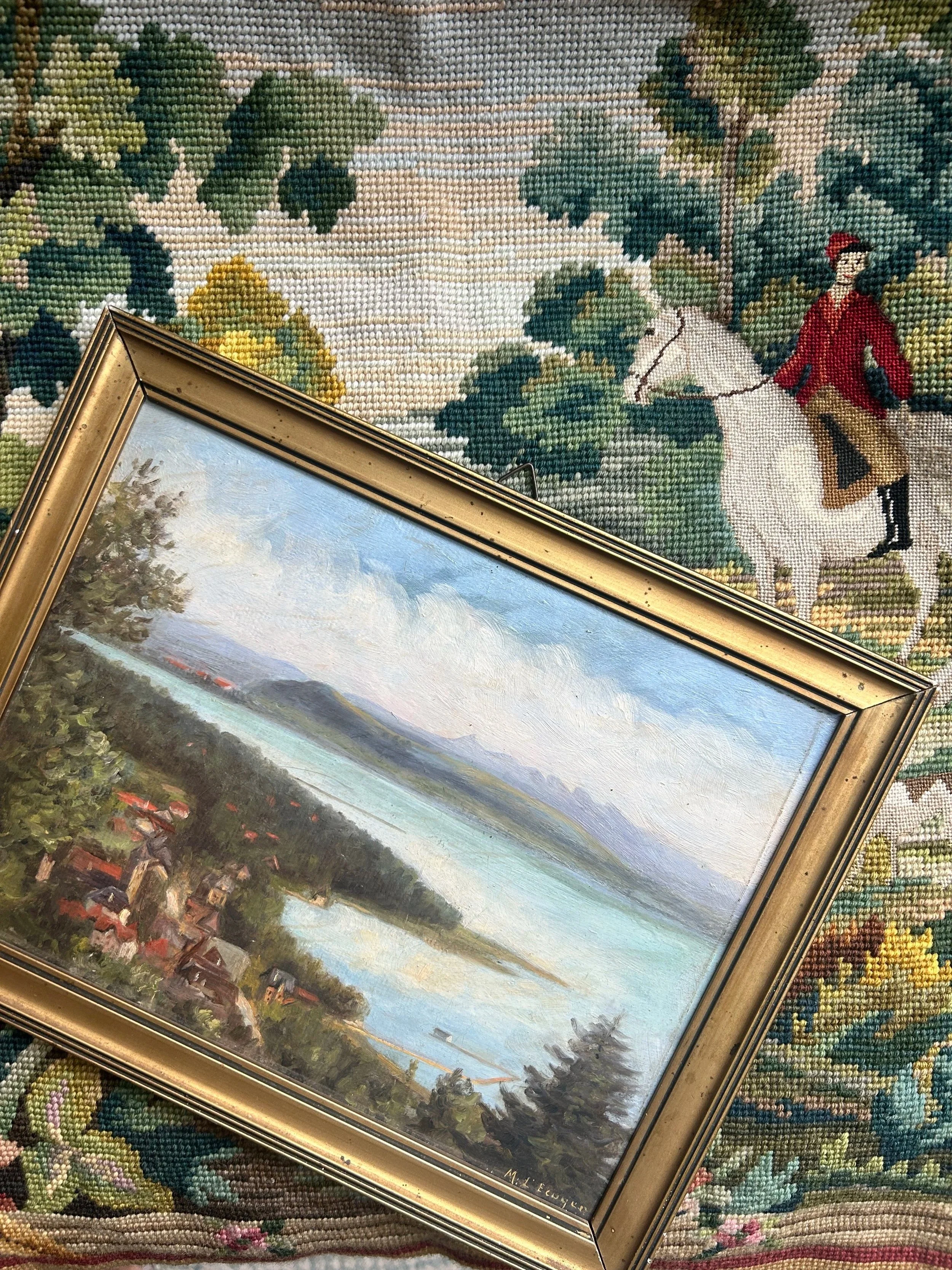 A landscape painting of a coastal scene with water, trees, and distant mountains, in a gold frame. A tapestry with a man riding a white horse amidst green trees is partially visible behind the painting.