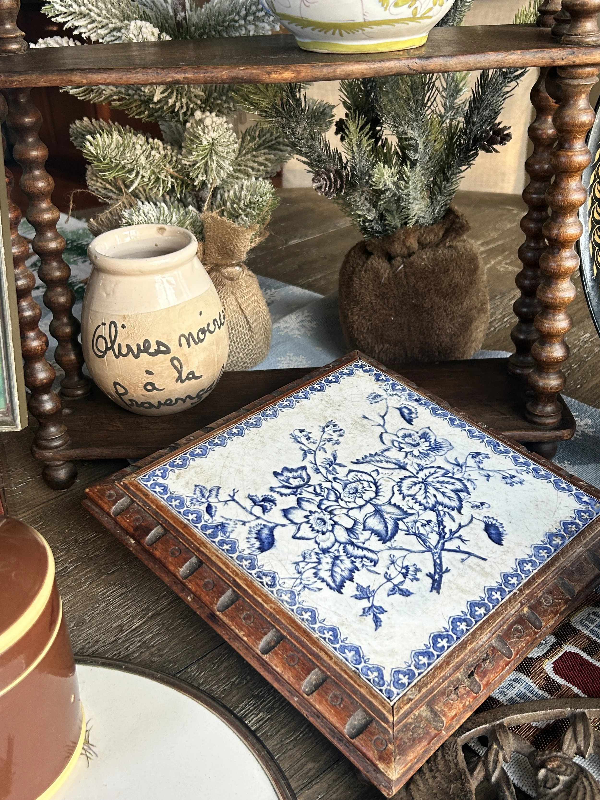 Decorative wooden shelf holding two small artificial Christmas trees in burlap and fuzzy wrapping, a cream-colored pot with French text, and a blue and white floral decorative tile on a dark wooden table.