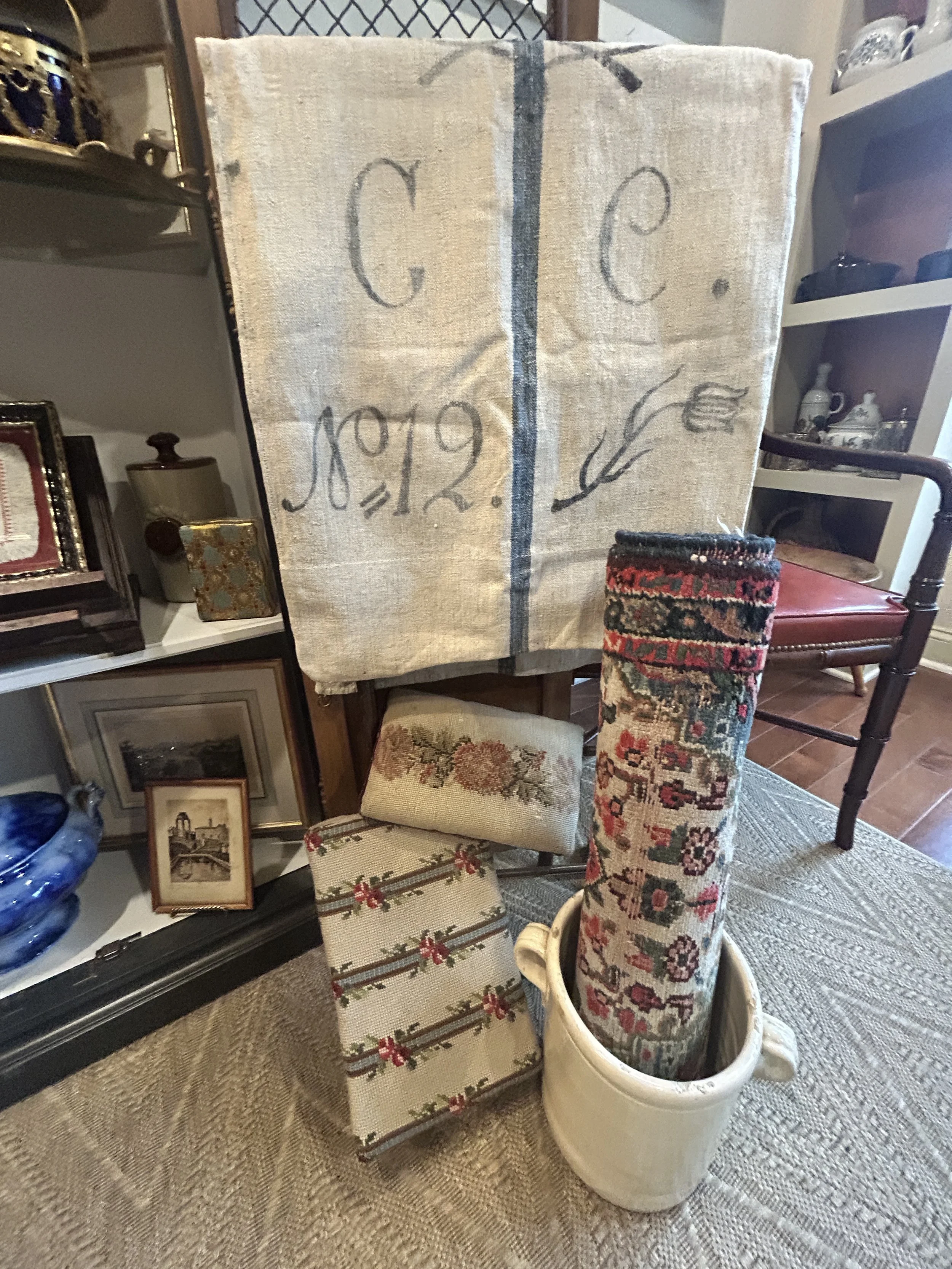 A collection of vintage items, including a woven banner with handwritten text, a rolled-up rug with floral patterns, a small upholstered stool with a floral design, and a white ceramic pot holding the rolled rug, all displayed against shelves with other decorative objects.