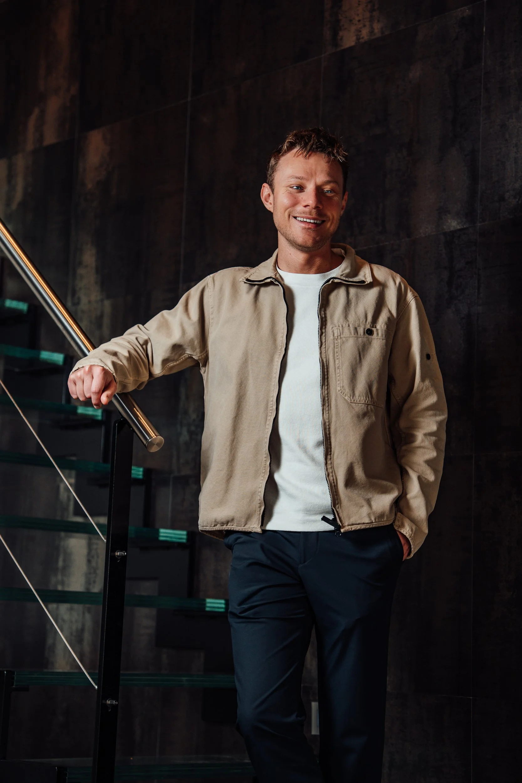 A smiling man wearing a beige jacket and white shirt standing next to a staircase with dark textured walls.