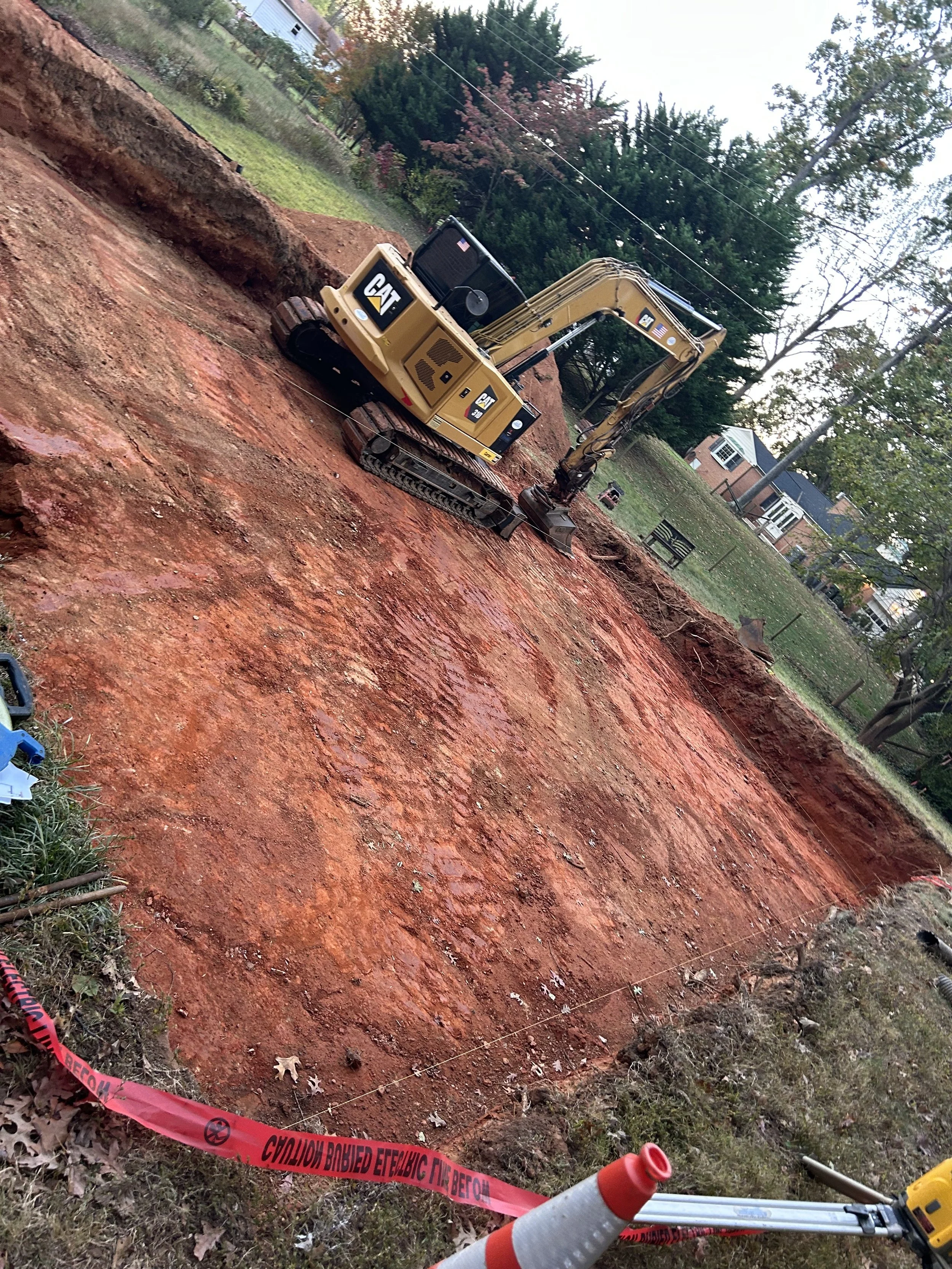 A construction site with a small yellow Caterpillar excavator digging into a large, uncovered red clay soil area. Residential houses and trees are visible in the background. Safety caution tape and an orange traffic cone are in the foreground.