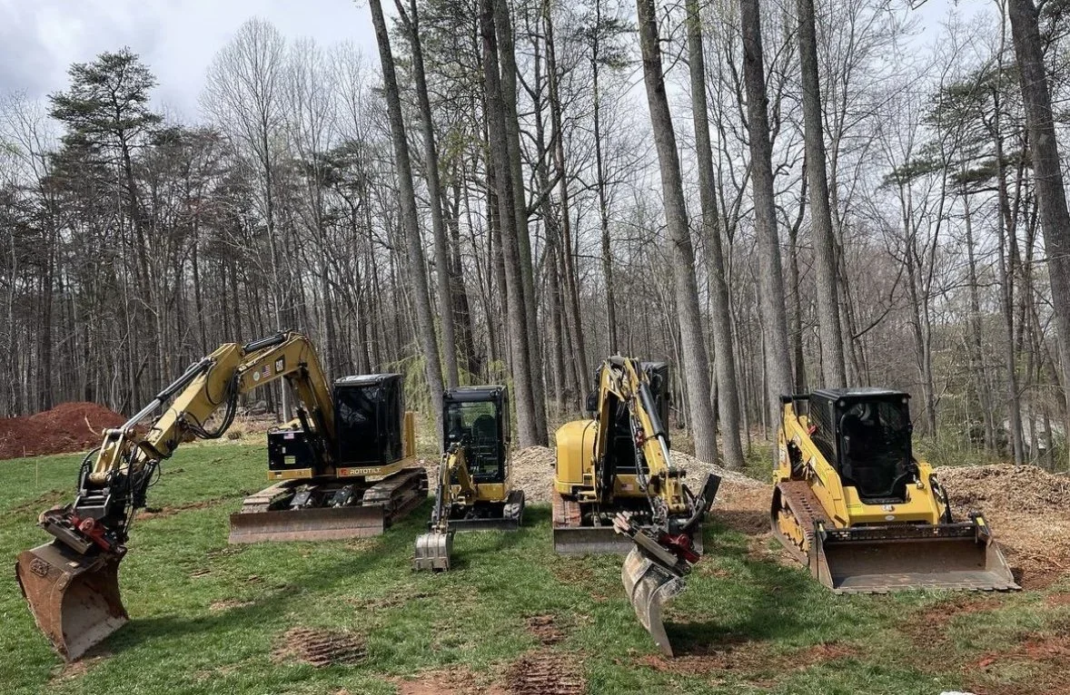 Four small yellow and black excavators parked on grass in front of a wooded area with leafless trees.