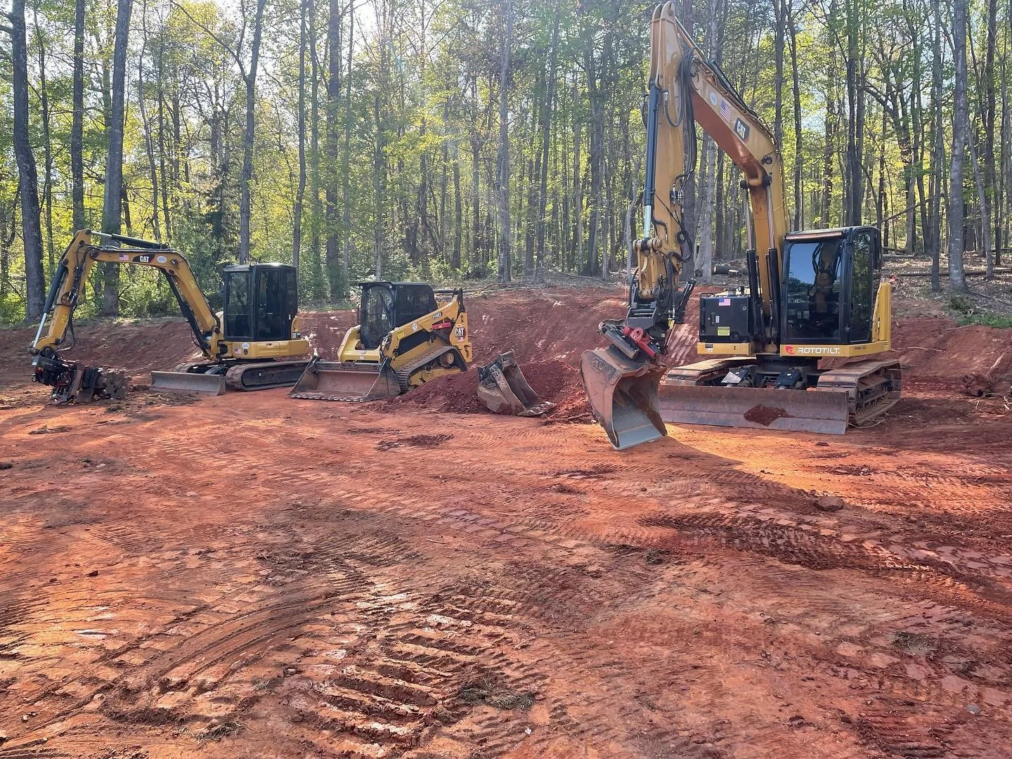 Three excavators working on a construction site with red soil and trees in the background.