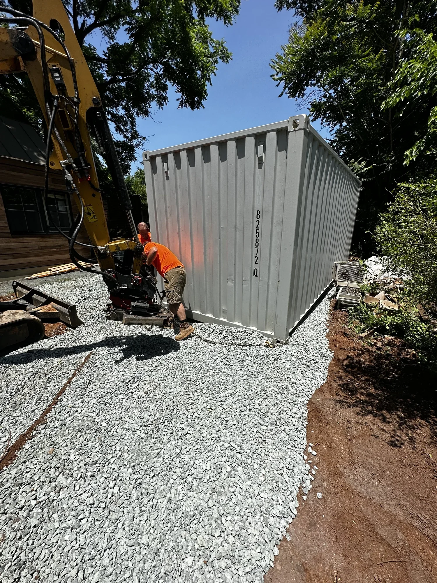 Worker in orange shirt and beige shorts using machinery to move a large metal shipping container in a yard with gravel, trees, and nearby building under a sunny sky.