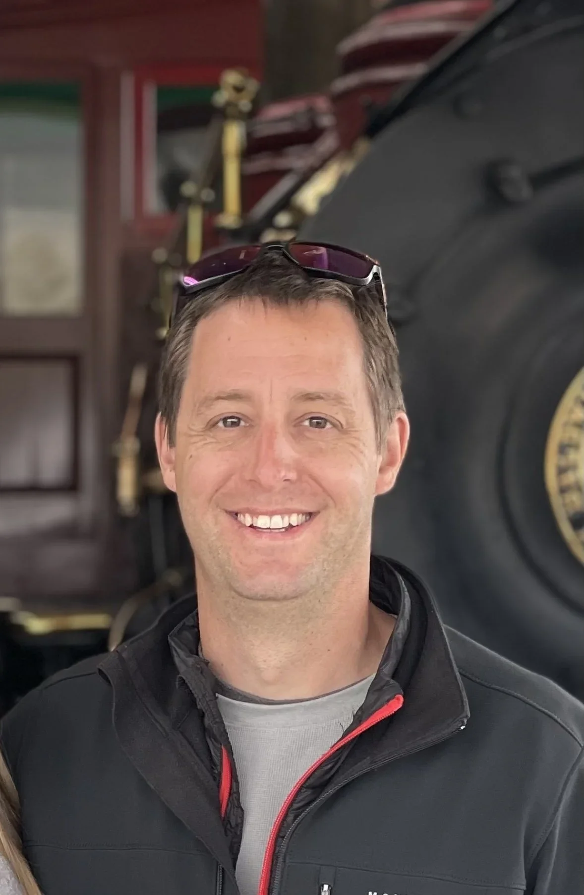 A man smiling, wearing a black jacket and gray shirt, with sunglasses on his head.