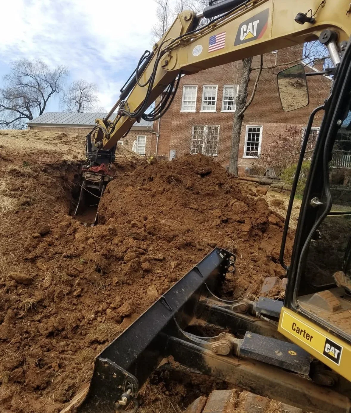A construction excavator with a yellow arm and black body digging a hole in the ground near a brick building with trees in the background.