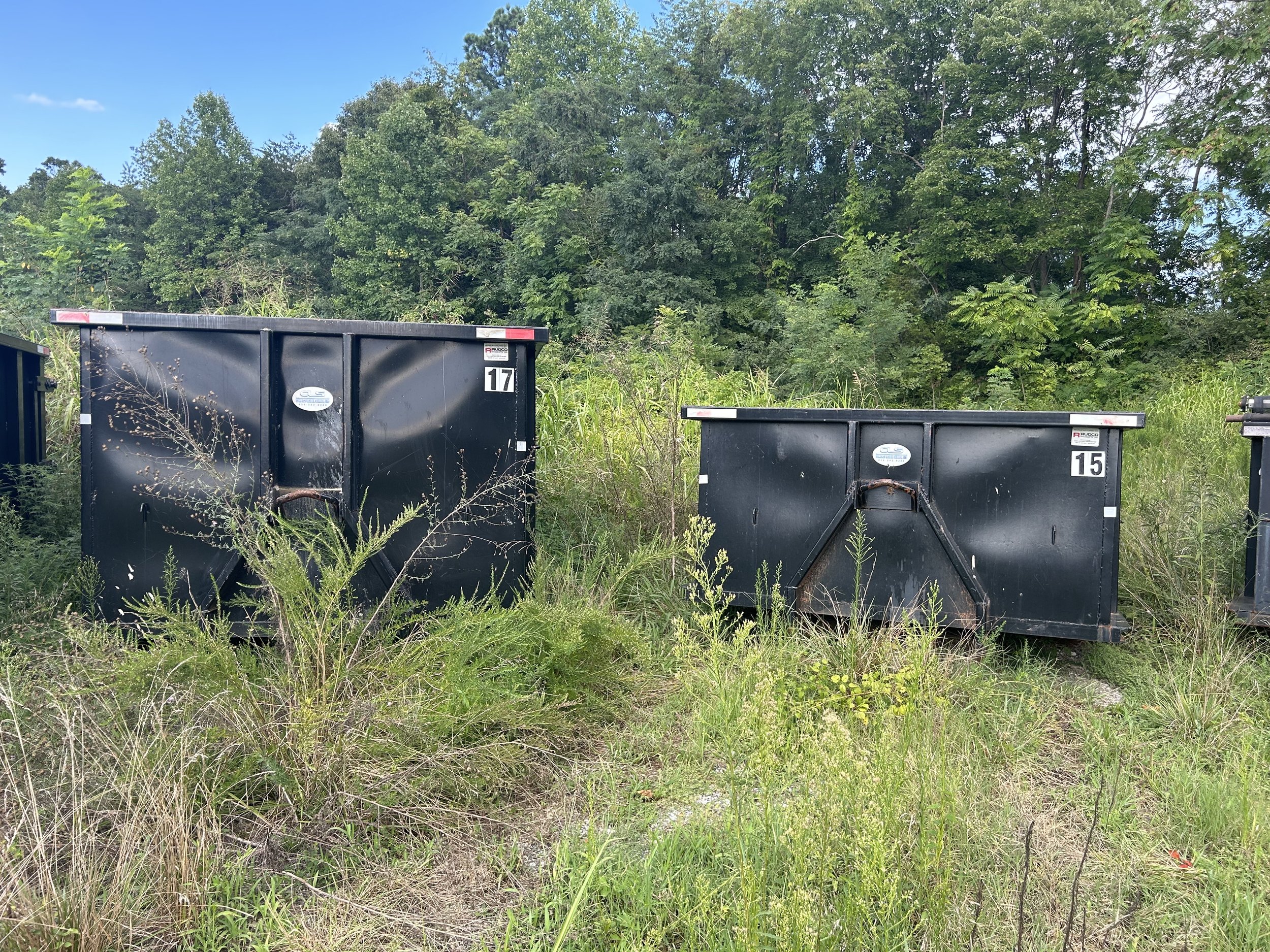 Two black trash containers numbered 17 and 15 in an overgrown grassy area with trees in the background.