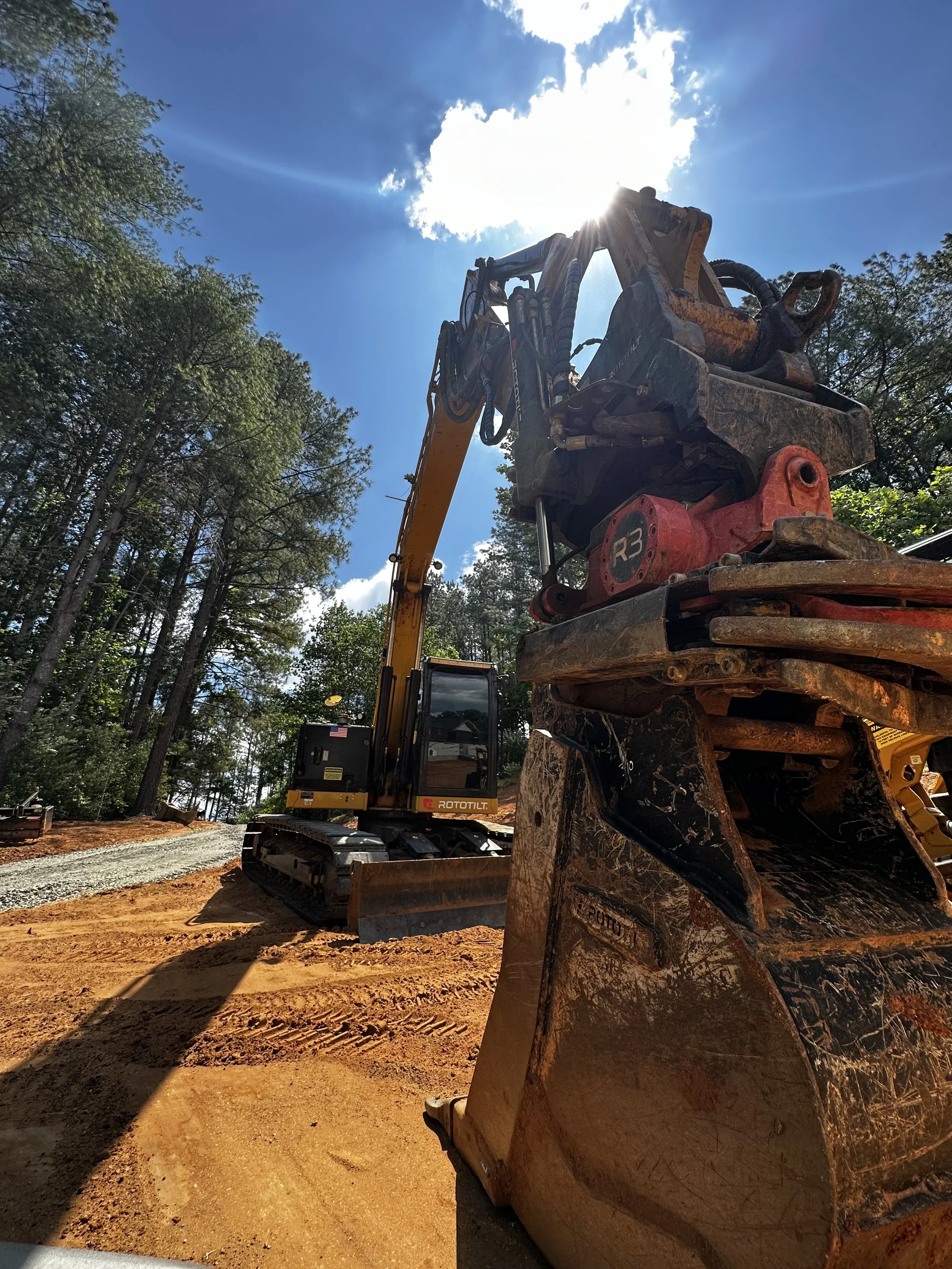 Construction excavator on dirt road under a bright sky with clouds, surrounded by trees.