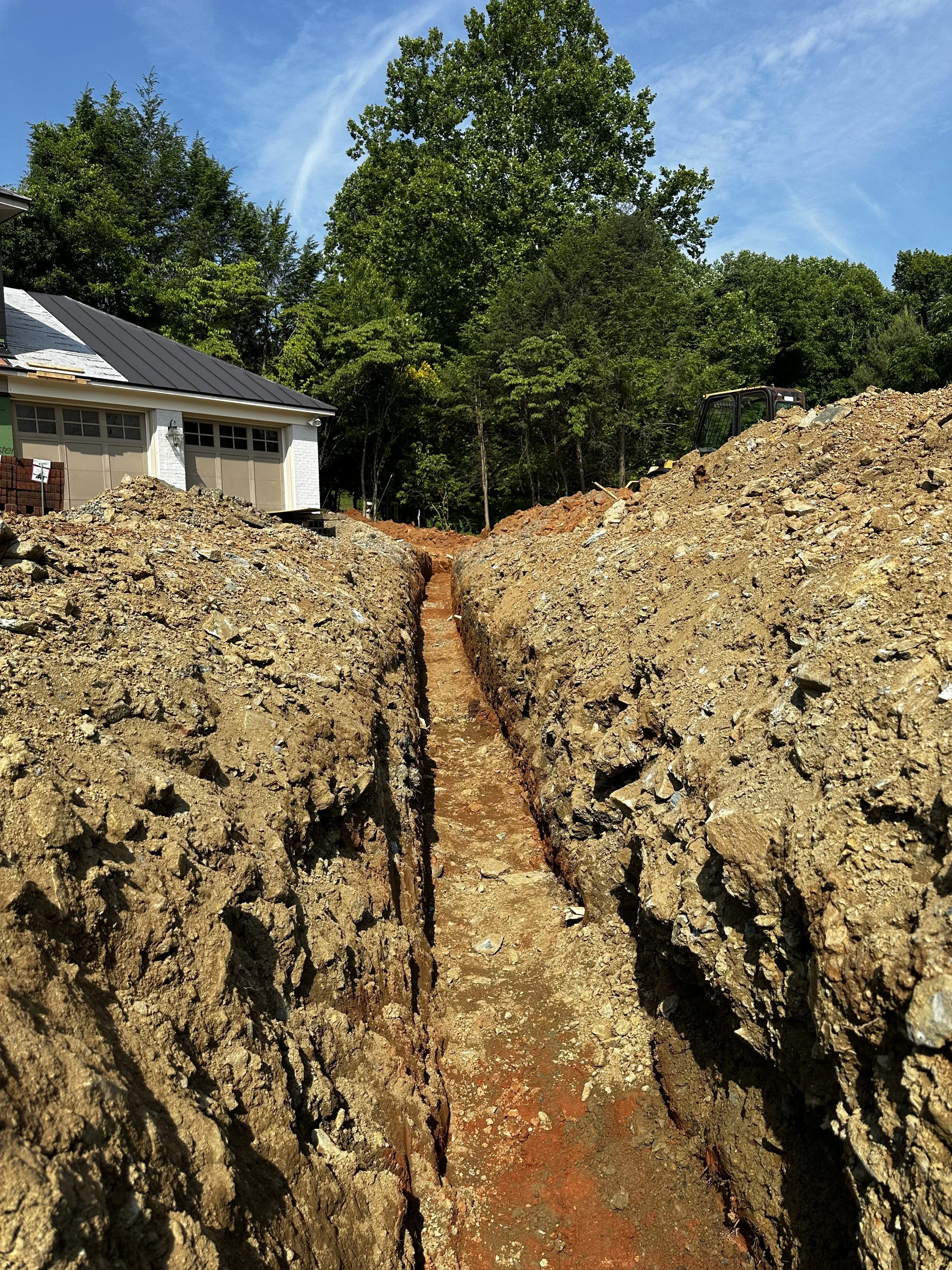 A construction site with a deep trench in the ground, dirt piles on either side, a house with a garage and lush green trees in the background, and a small excavator on the right side.