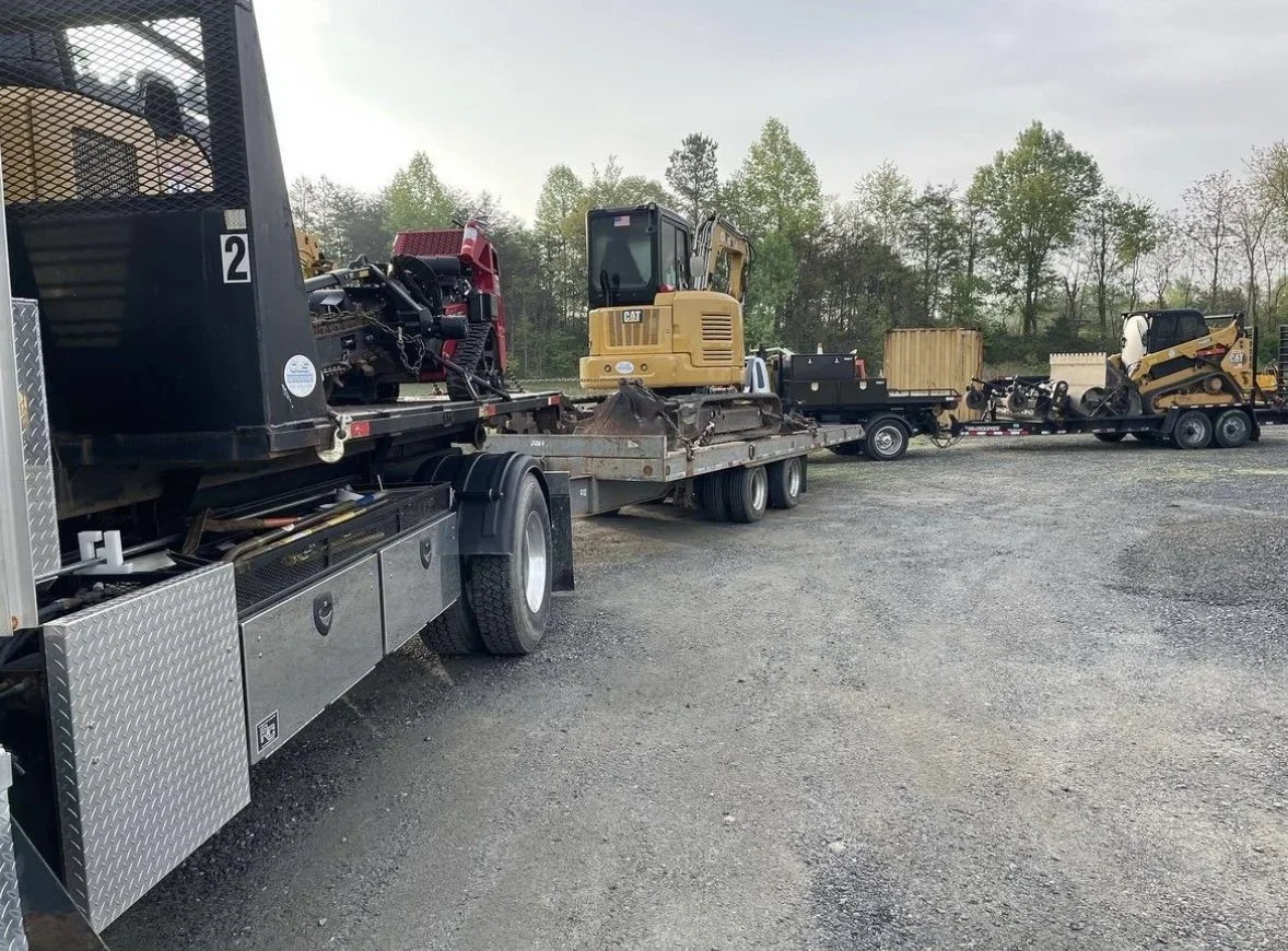 Various construction equipment and machinery loaded on trailers in an outdoor lot with trees in the background.