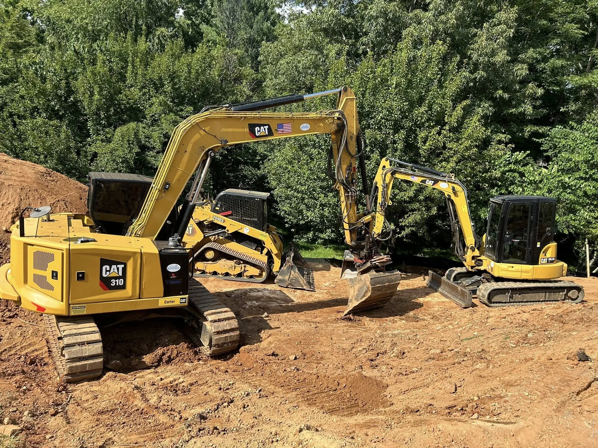 Two yellow Caterpillar excavators working on a dirt construction site near green trees.