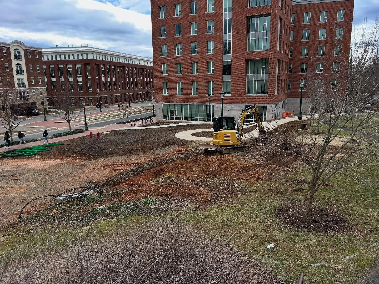 Construction site with a yellow excavator digging a trench in a grassy area near modern brick apartment buildings, with pedestrians walking on the sidewalk in the background.
