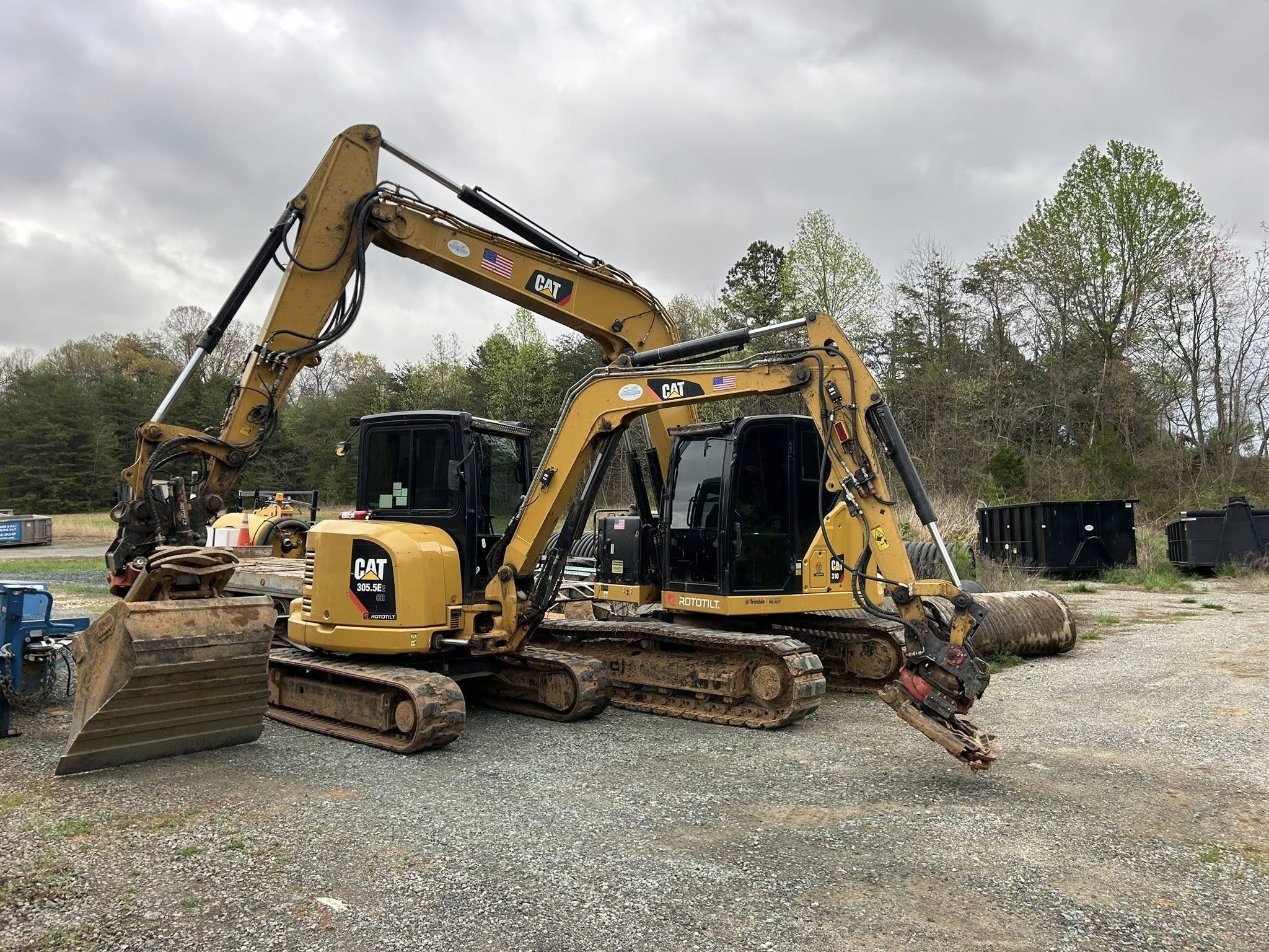Two yellow Caterpillar excavators parked on gravel with trees and cloudy sky in the background.