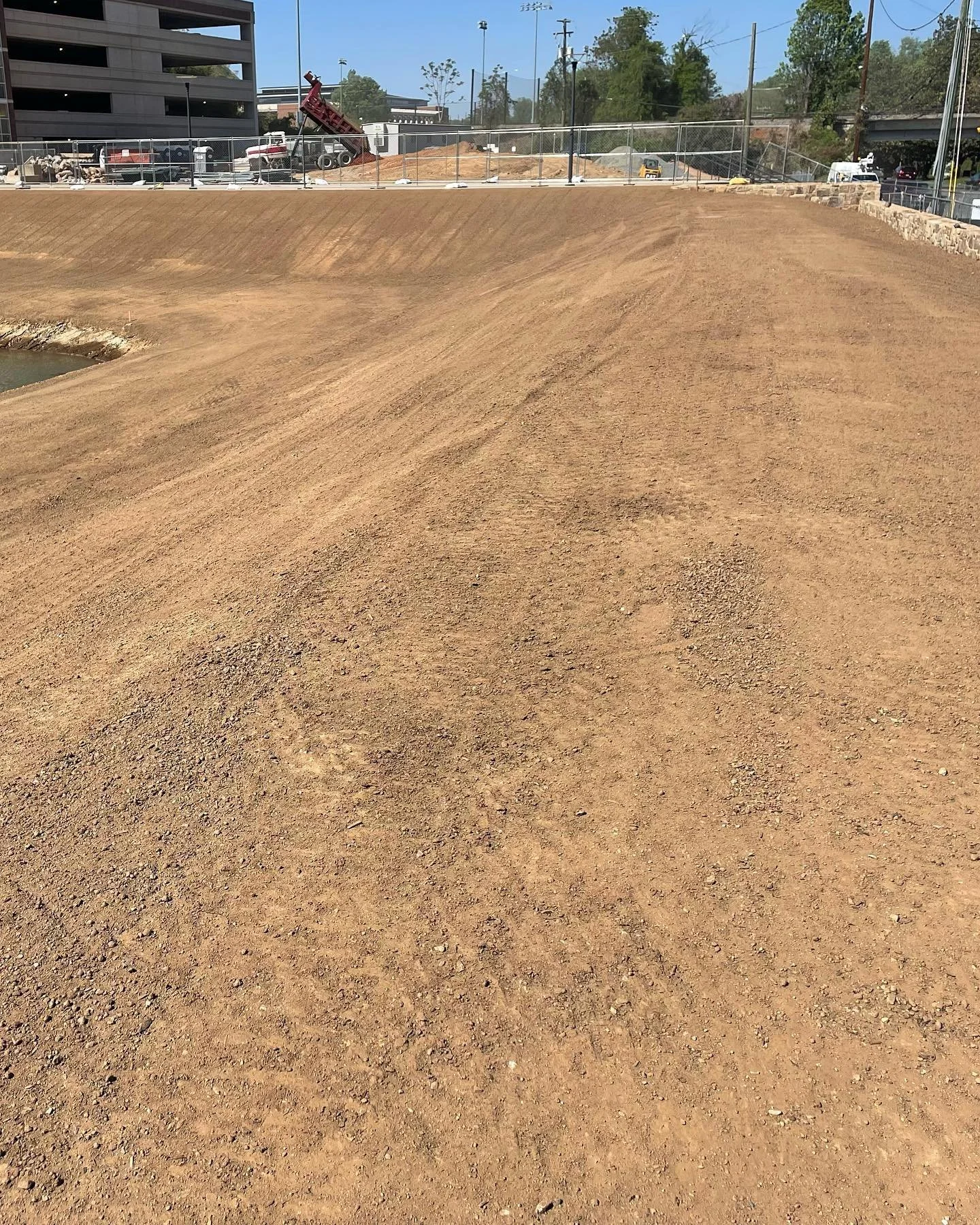 A construction site with dirt ground, a small pond on the left, and a chain-link fence in the background. There are construction vehicles and buildings in the distance.
