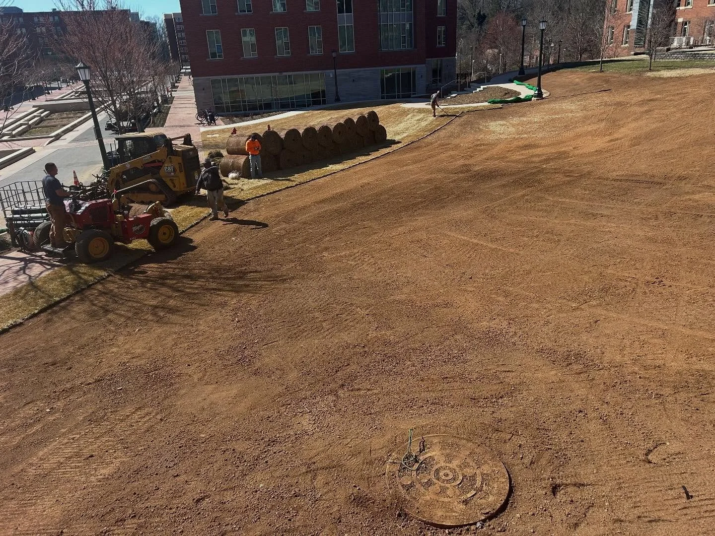 Construction workers leveling and preparing the ground on a hillside, with a small excavator, hay bales, and a green pipe, near a pathway with brick buildings and leafless trees in the background.