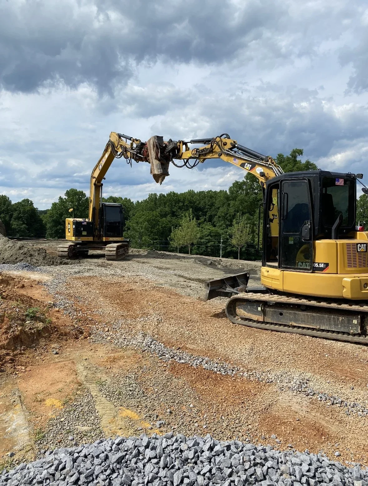 Two yellow Caterpillar excavators working on a construction site with gravel and dirt, surrounded by trees and a cloudy sky.