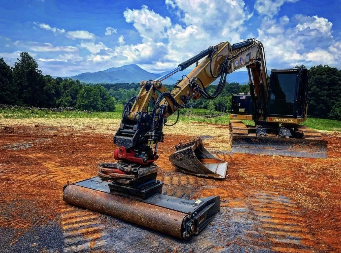 A yellow excavator operating on a dirt lot with a mountain and trees in the background under a blue sky with clouds.