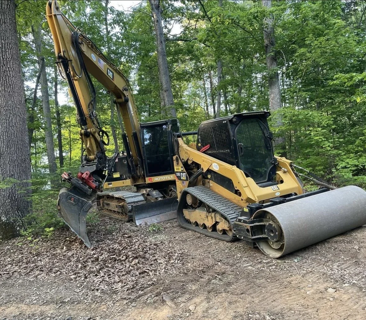 A construction roller machine with a yellow and black color scheme is parked on a dirt path in a wooded area, surrounded by trees.