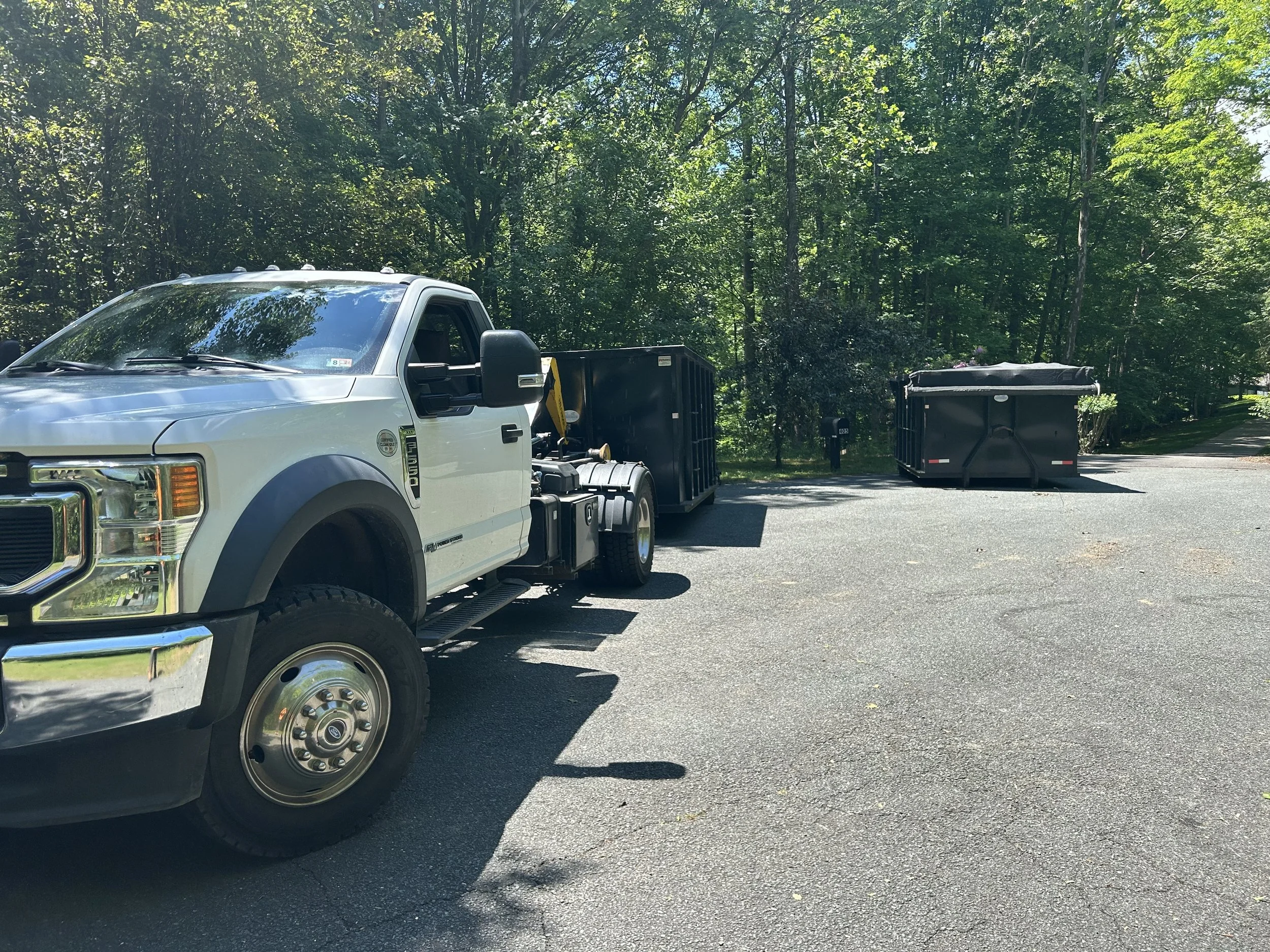 A white pickup truck with an attached black waste container is parked on a driveway, with two large black trash bins in the background surrounded by green trees under a clear blue sky.