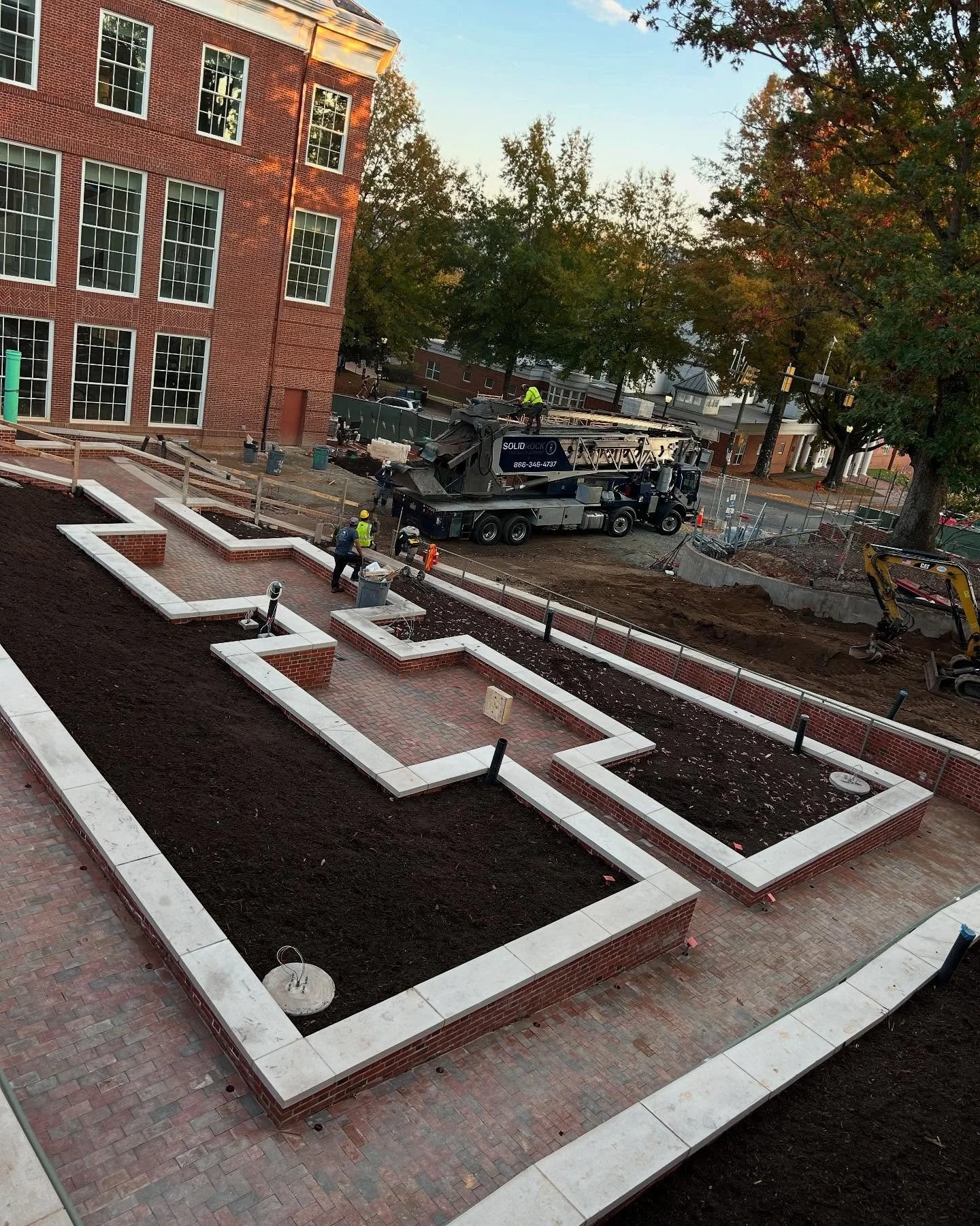Construction workers working on landscaping and brickwork at a building site, with a crane vehicle, trees, and a brick building in the background during daylight.