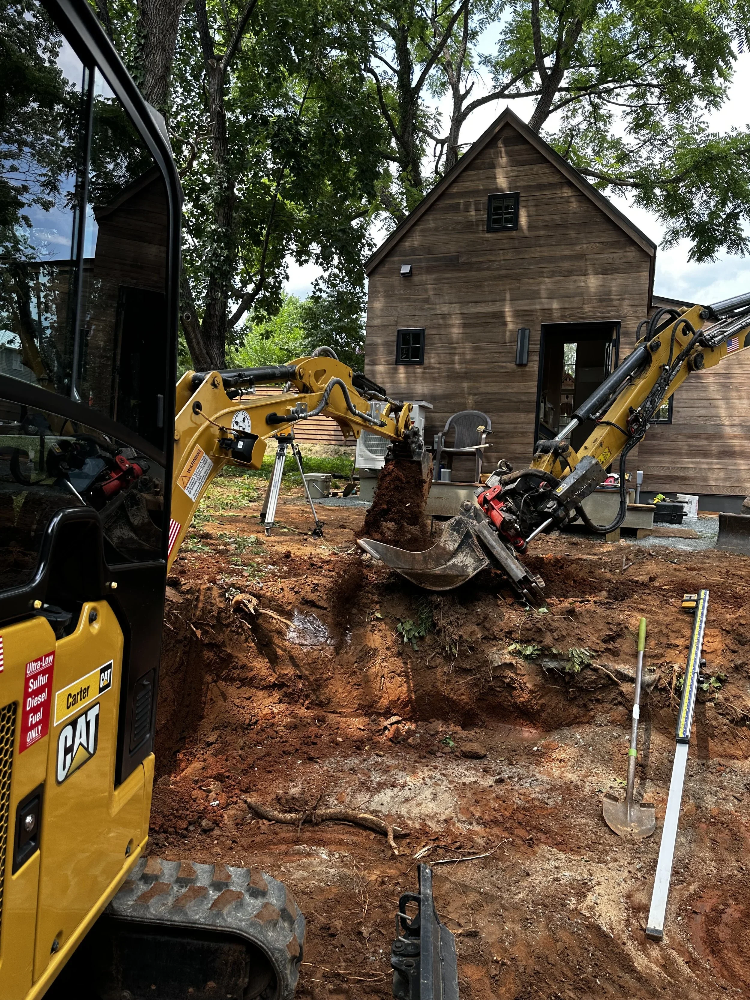 Construction site with a yellow excavator digging a trench in front of a wooden house surrounded by trees.