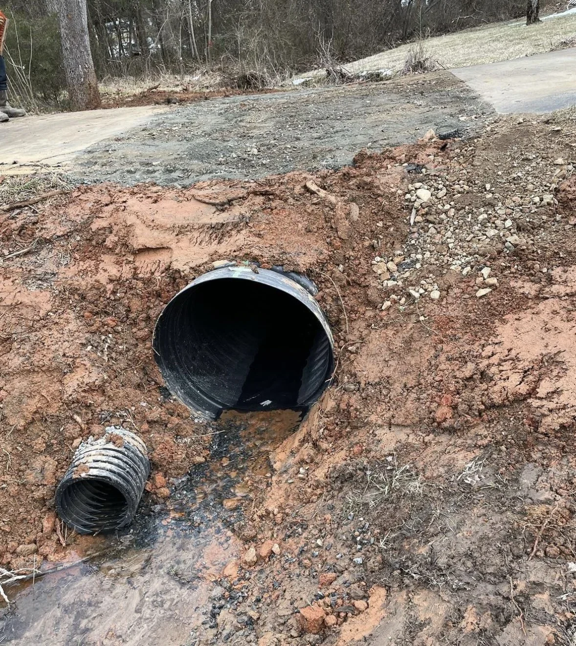 Underground construction site showing a large black piping pipe with a smaller pipe nearby, surrounded by dirt and soil, with a forest and road in the background.