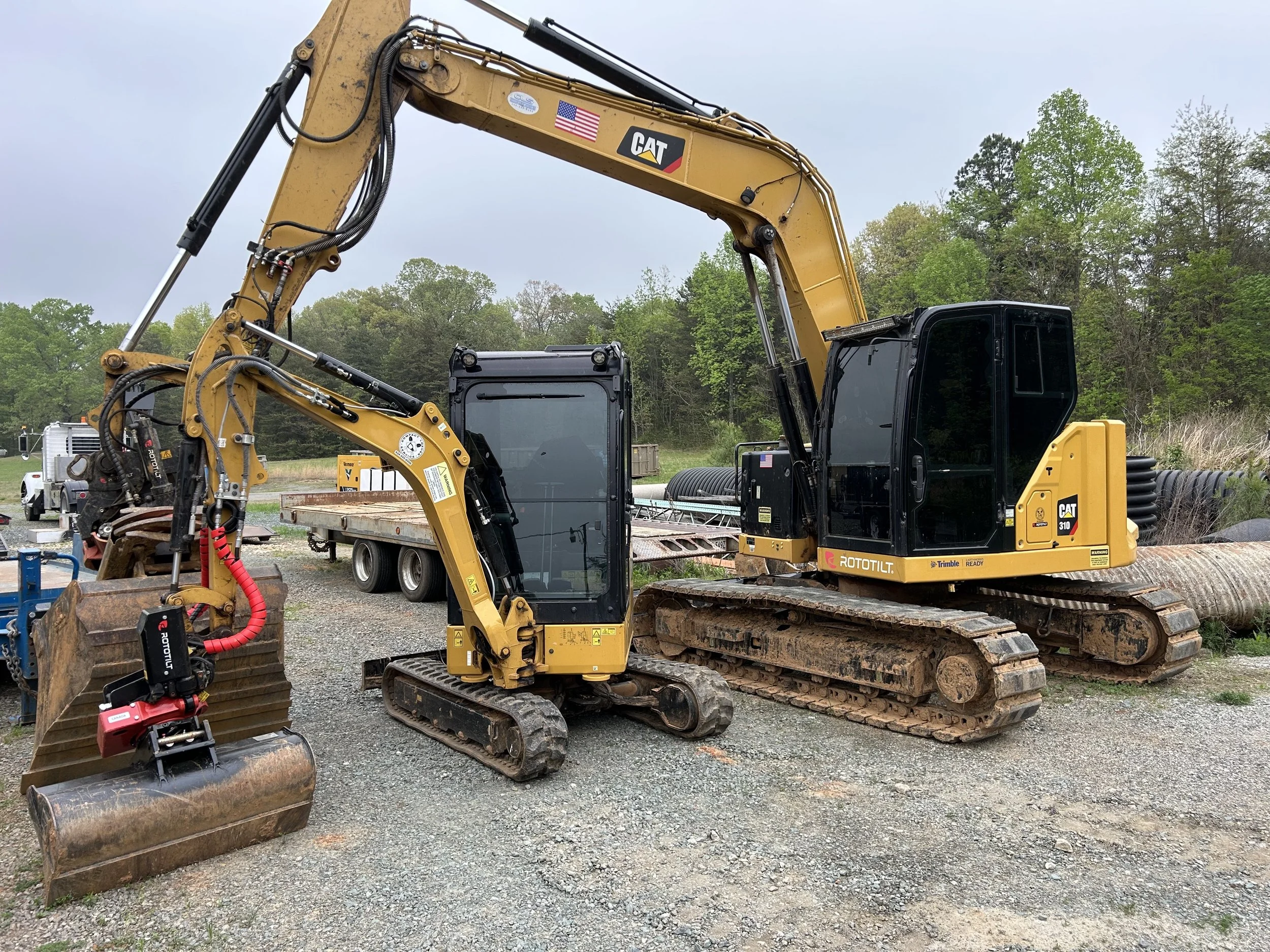 A large yellow Caterpillar excavator on a construction site, equipped with a bucket attachment, positioned on gravel ground with trees in the background.