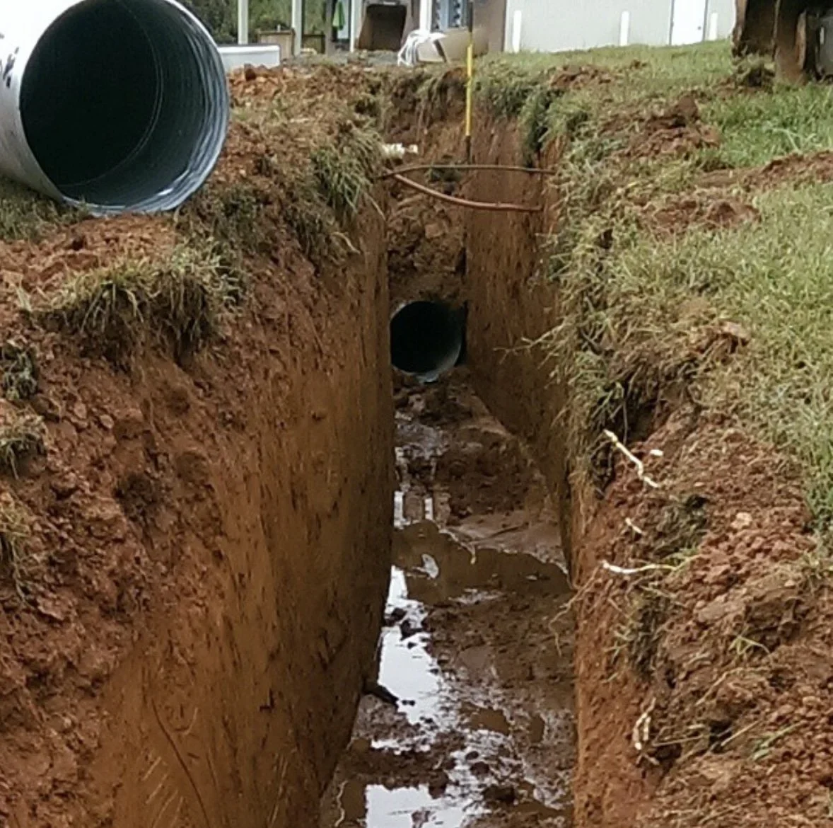 A trench with a large pipe installed, surrounded by dirt and grass, with another pipe laying on the ground at the edge of the trench.