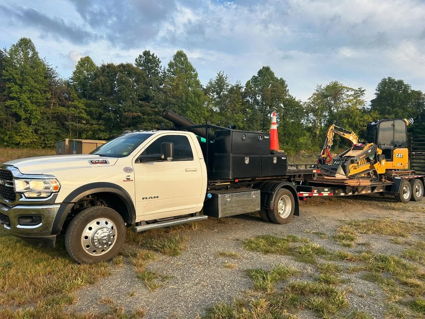 A white RAM pickup truck with a flatbed carrying construction equipment, including a small excavator, and safety cones, parked on a gravel lot with trees in the background.