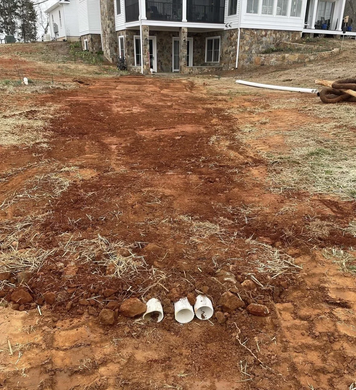 A construction site in front of a house with reddish dirt, three white PVC pipes near the foreground, and some construction materials to the right. The house has a stone foundation and white siding.