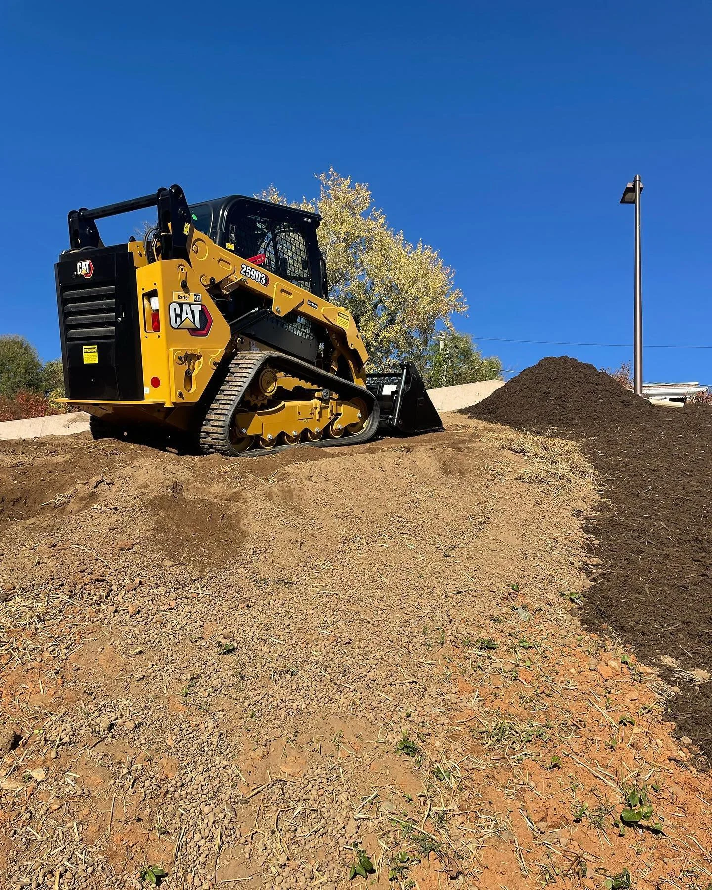 A small yellow and black Caterpillar compact excavator on a dirt construction site with a mound of soil and a clear blue sky.