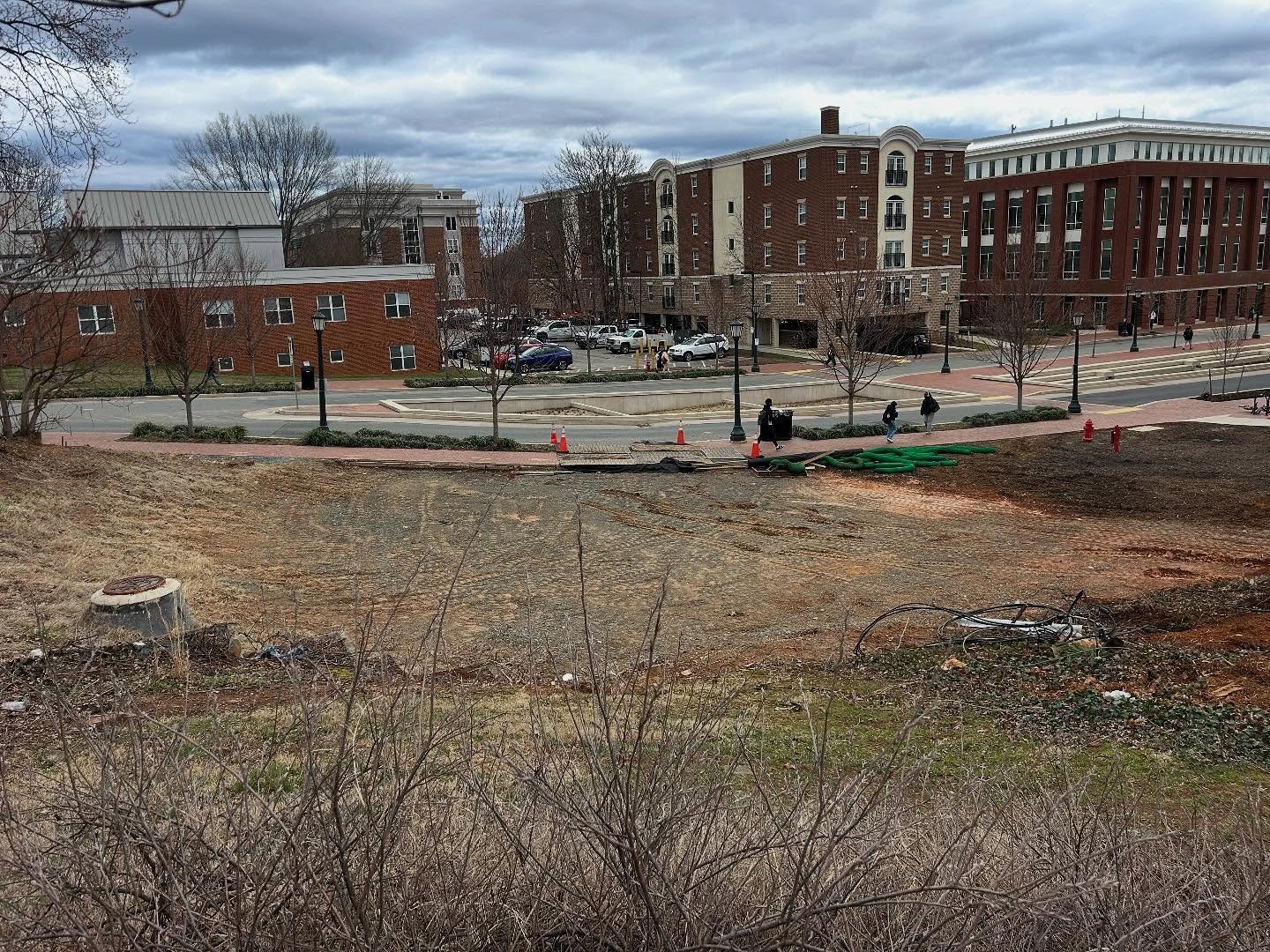 Construction site with a cleared dirt area in the foreground, urban buildings in the background, and a walkway with pedestrians.