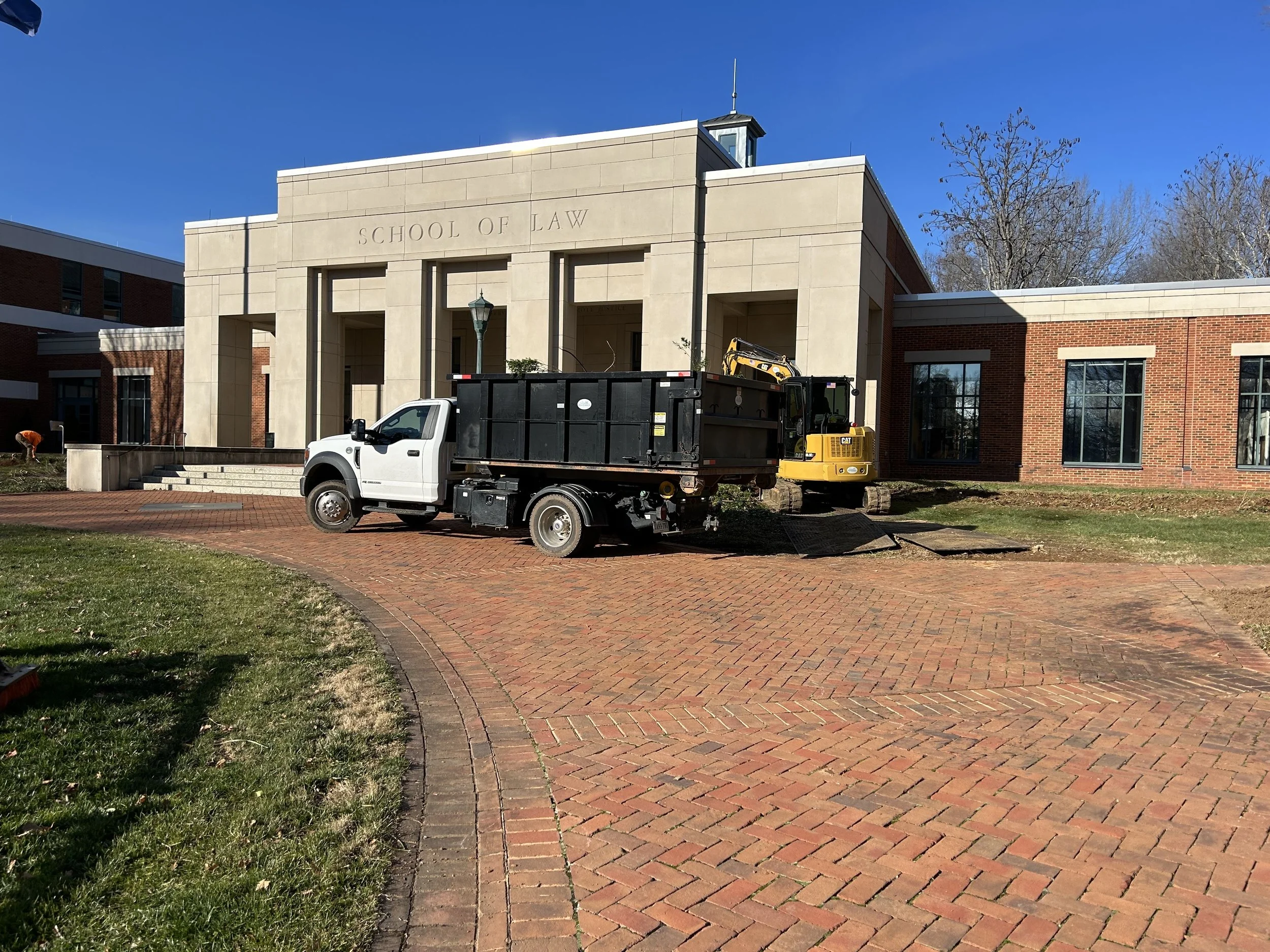 Construction equipment, including a small excavator and a pickup truck, outside a school of law building with red brick and beige stone facade.