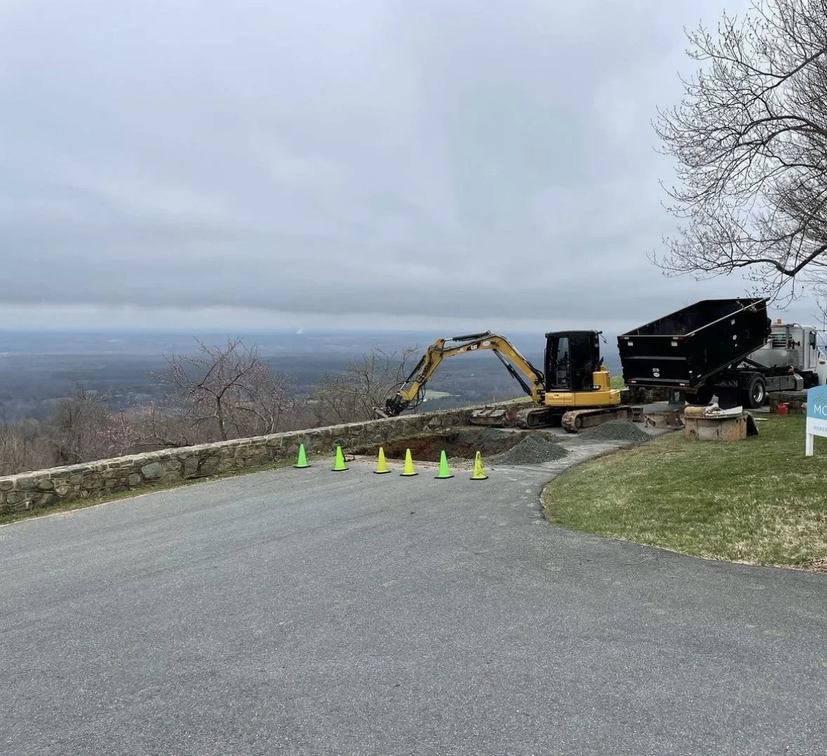 Construction site on a hill with a small excavator, a dump truck, and construction cones, overlooking a scenic landscape with a cloudy sky.